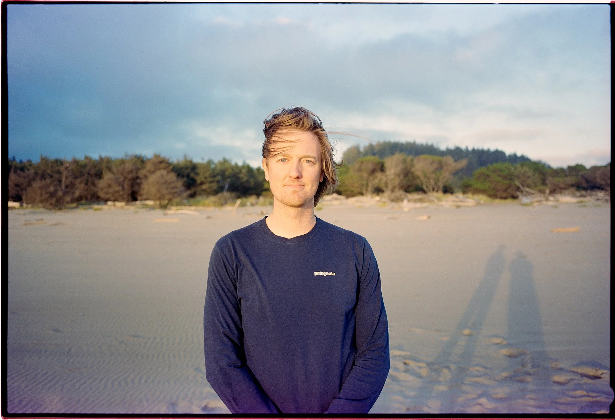 A young man with light brown hair in a black Patagonia shirt standing on a sandy beach with trees and hills in the background, during late afternoon or early evening.