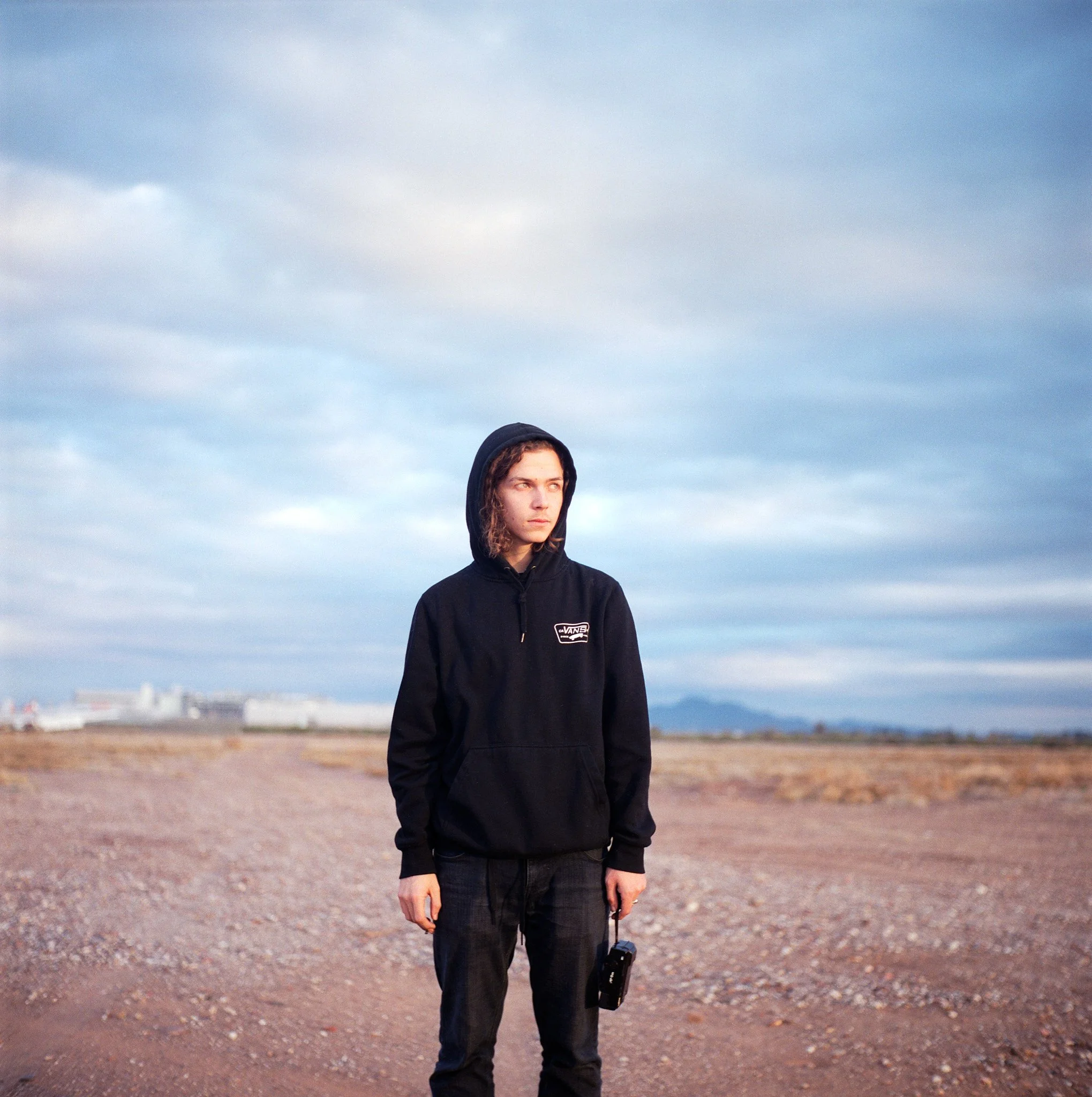 A young woman with curly hair wearing a black hoodie with a 'Vans' logo, standing on a dirt path in a flat, open landscape under a cloudy sky, holding a small black camera or device in her right hand.