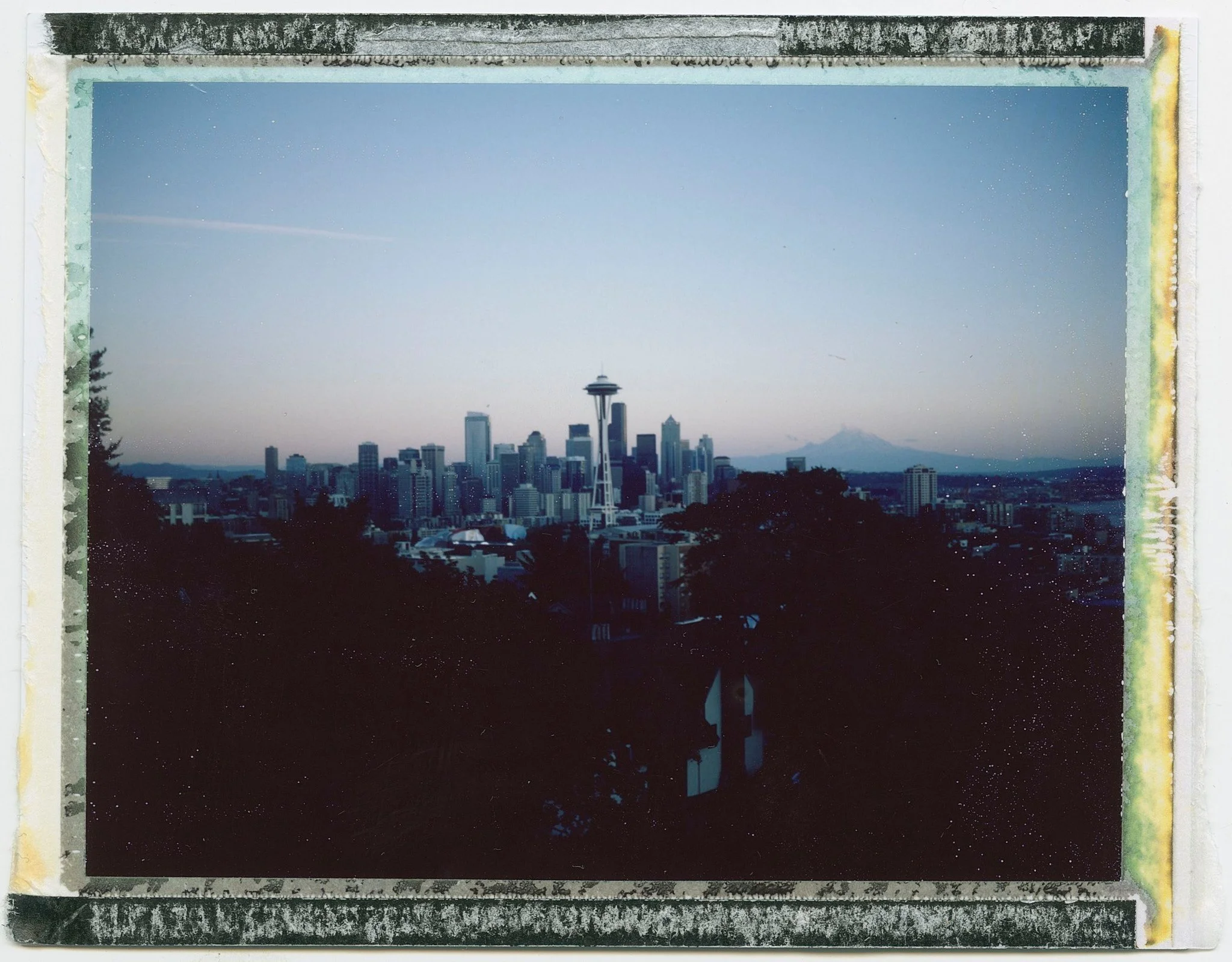 A photograph of the Seattle skyline with the Space Needle in the center, taken at dusk or dawn, with trees in the foreground and mountains in the background.