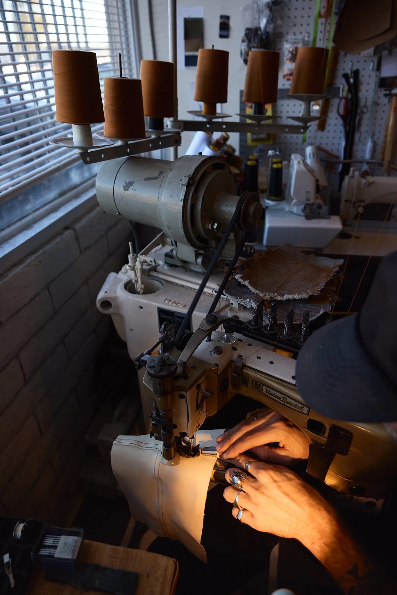 Person sewing fabric on a vintage industrial sewing machine in a workshop, with spools of thread above and tools in the background.