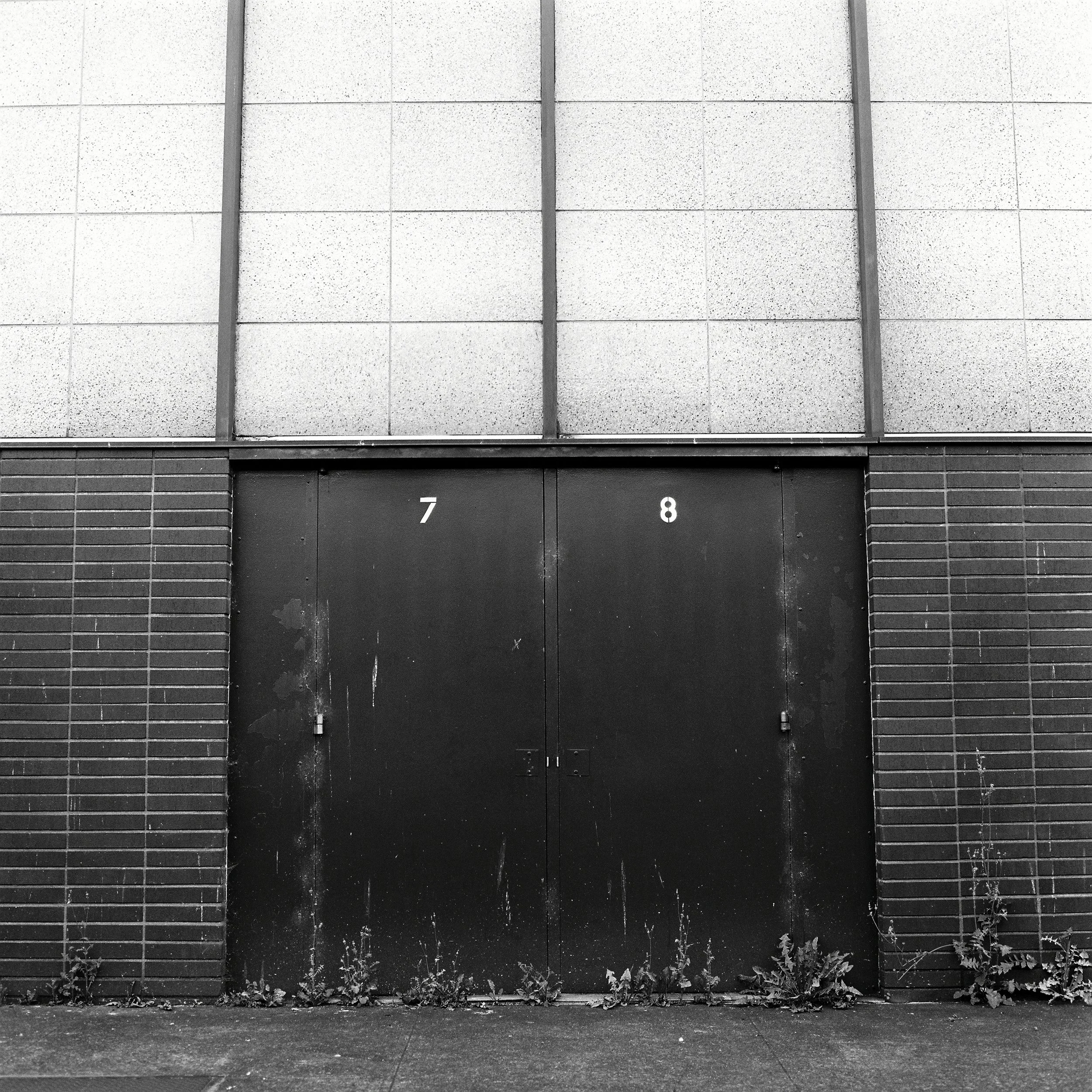 Black metal door with the numbers 7 and 8 above it, on a brick and concrete building, with weeds growing at the base.