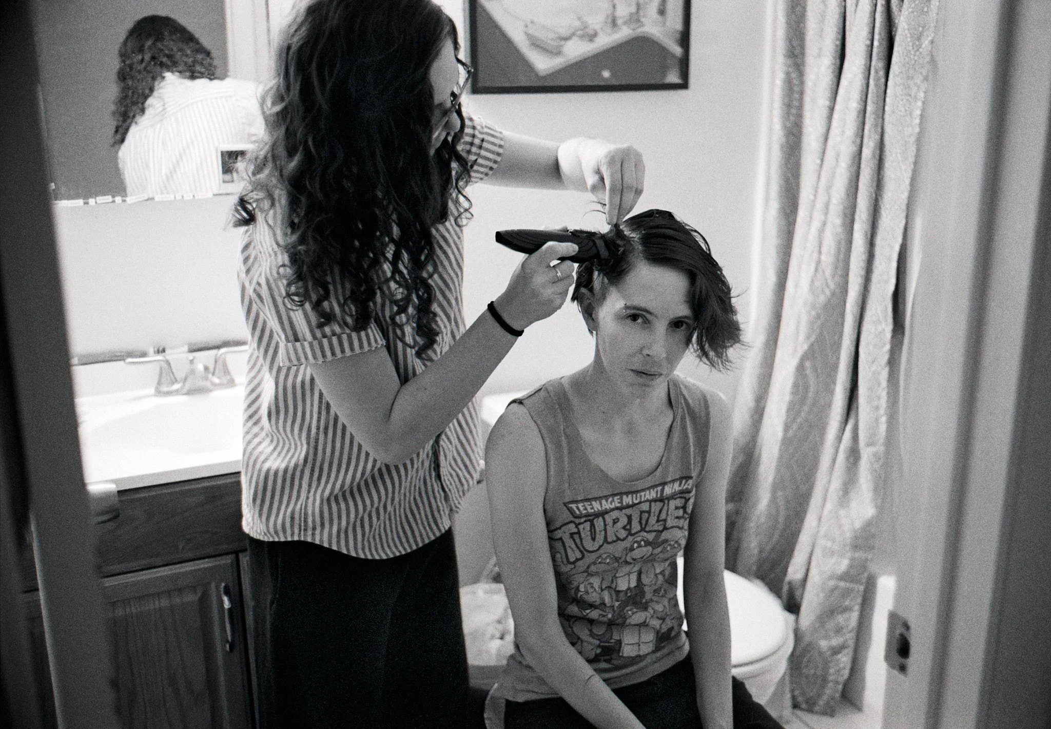 A woman sitting on a bathroom counter getting her hair styled by another woman. The stylist uses a curling iron on the seated woman's hair. A mirror reflects the stylist, and a framed picture hangs on the wall behind them. There are bathroom curtains