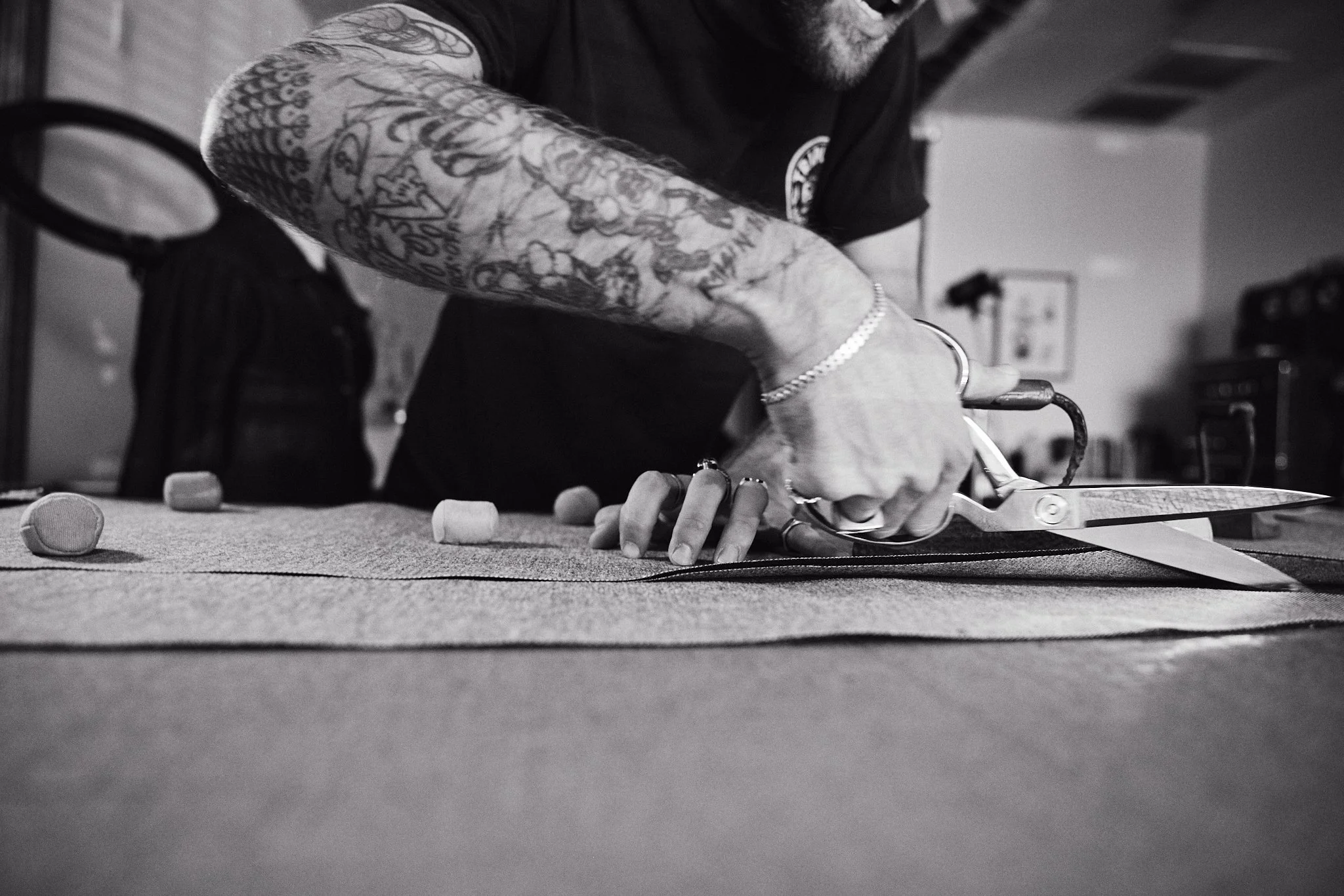 Close-up black and white photo of a person with tattoos on their right arm cutting fabric on a table with scissors.