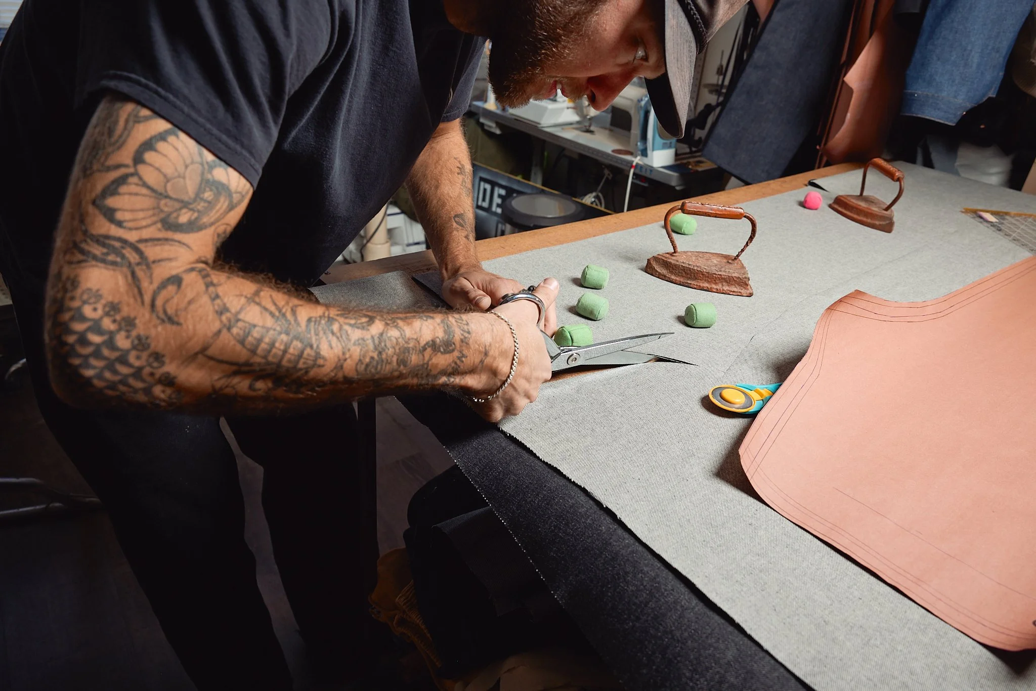A man with tattoos on his arm is cutting leather on a worktable. Several pieces of leather, sewing pattern paper, green foam pieces, and metal tools are on the table, along with a pair of scissors.