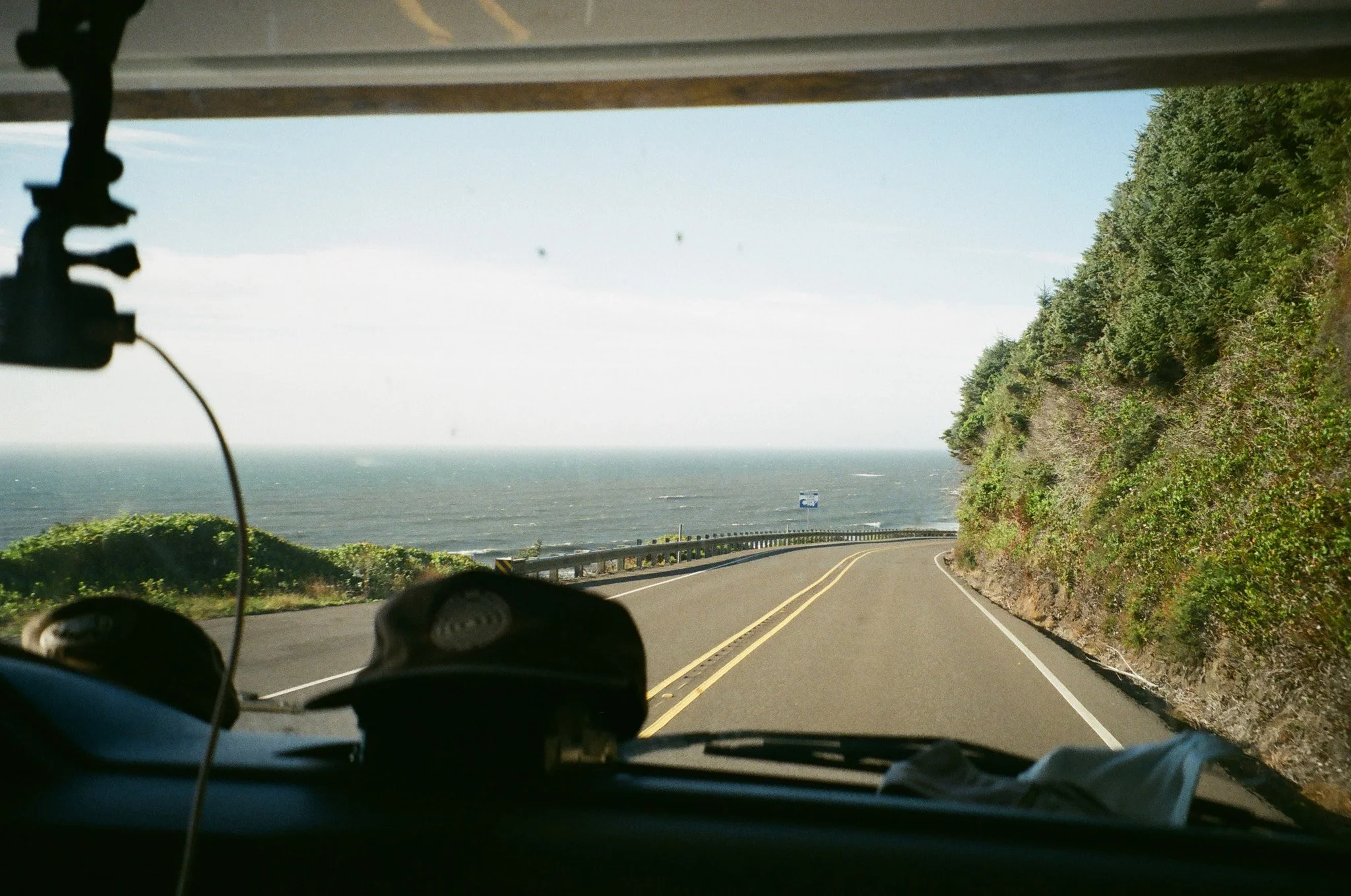 View from inside a vehicle driving along a coastal road with the ocean on the left and a hillside covered in green trees on the right.
