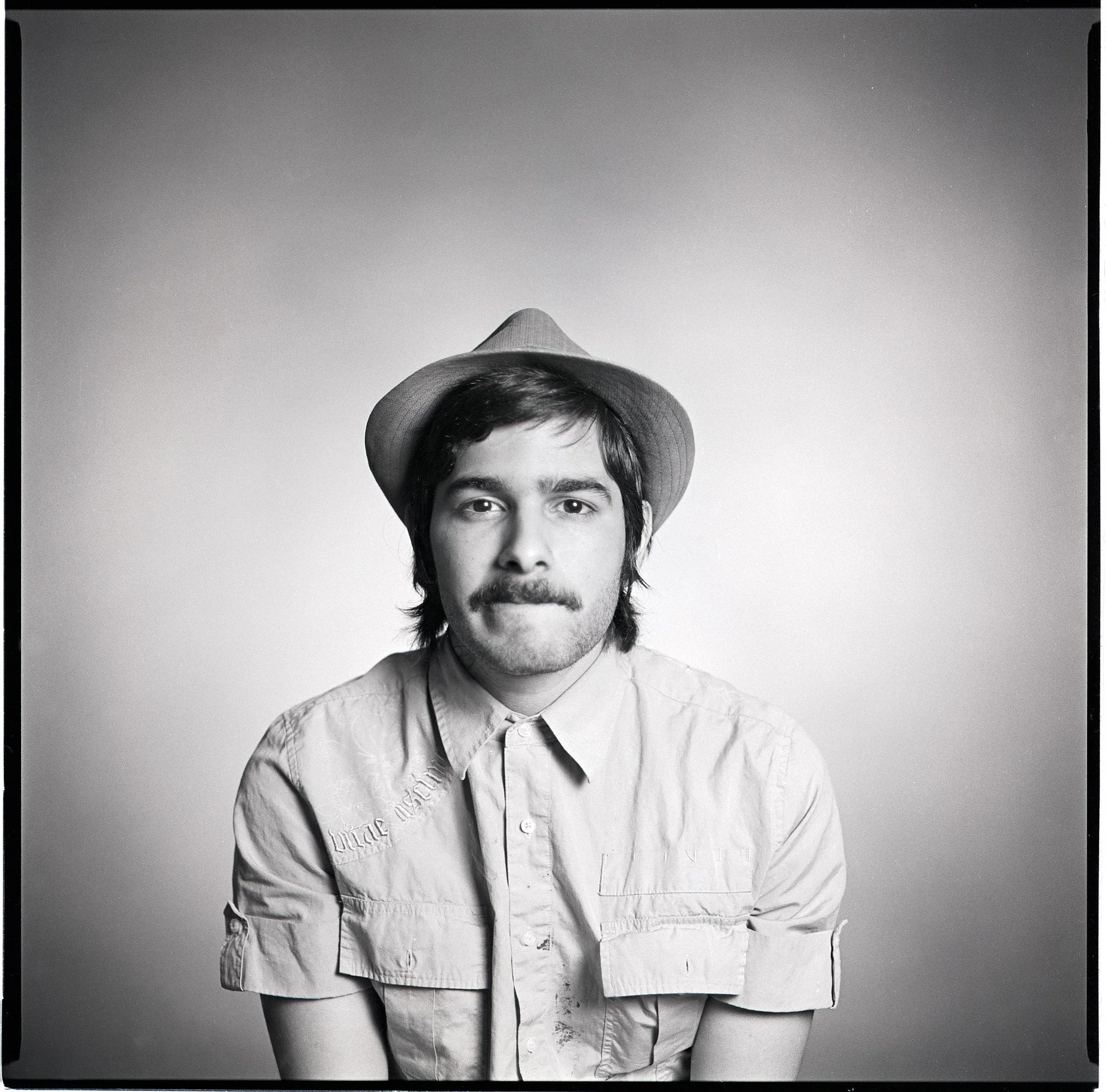 Black and white portrait of a man with a mustache and beard, wearing a light-colored short-sleeve shirt and a cowboy hat, facing forward against a plain background.