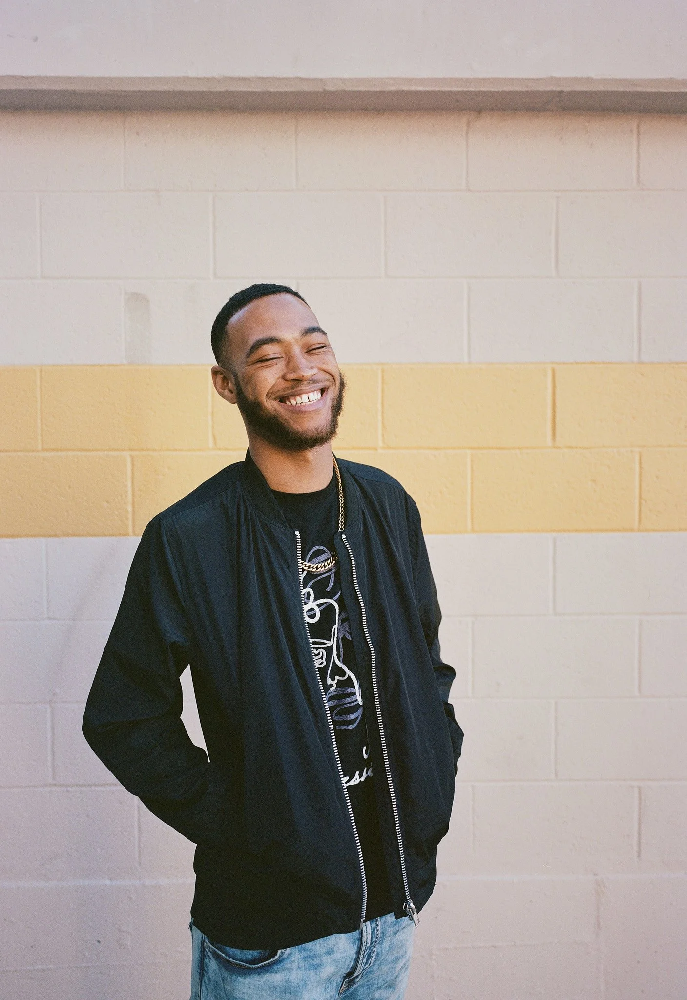 Smiling young man standing against a beige and yellow painted brick wall, wearing a black jacket and jeans.