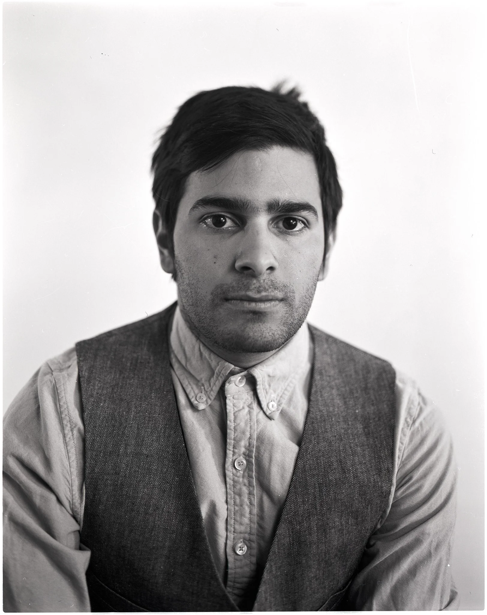 Black and white portrait of a young man with dark hair, wearing a button-up shirt and a vest, looking directly at the camera.