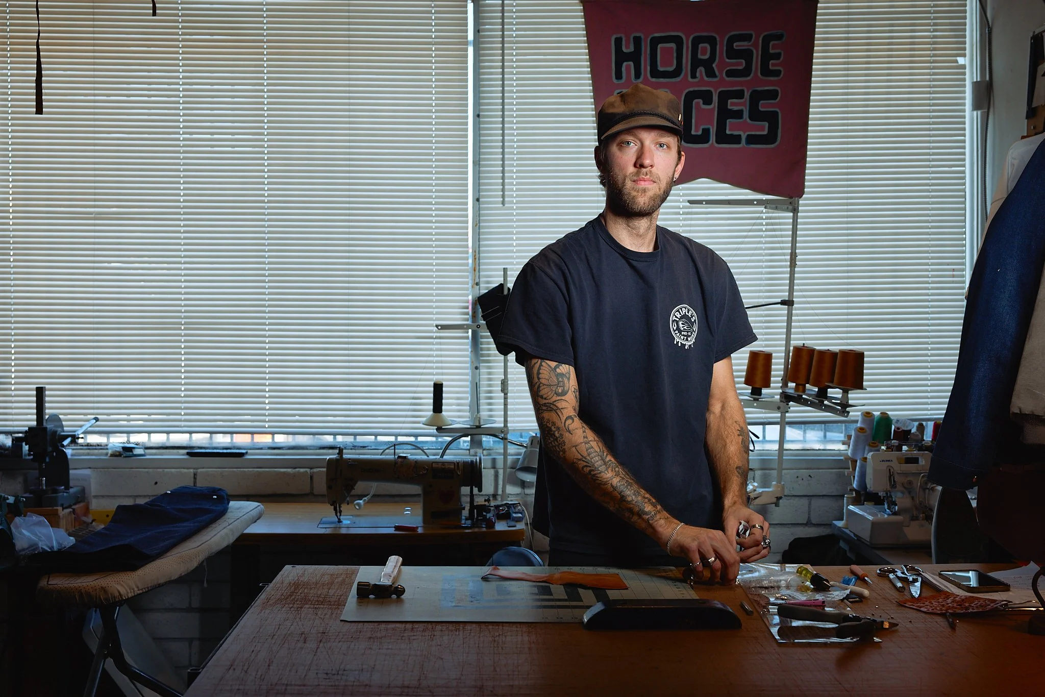 A man with tattoos standing in a workshop, surrounded by sewing and crafting materials, with a backdrop of blinds and a 'Horse Faces' banner.