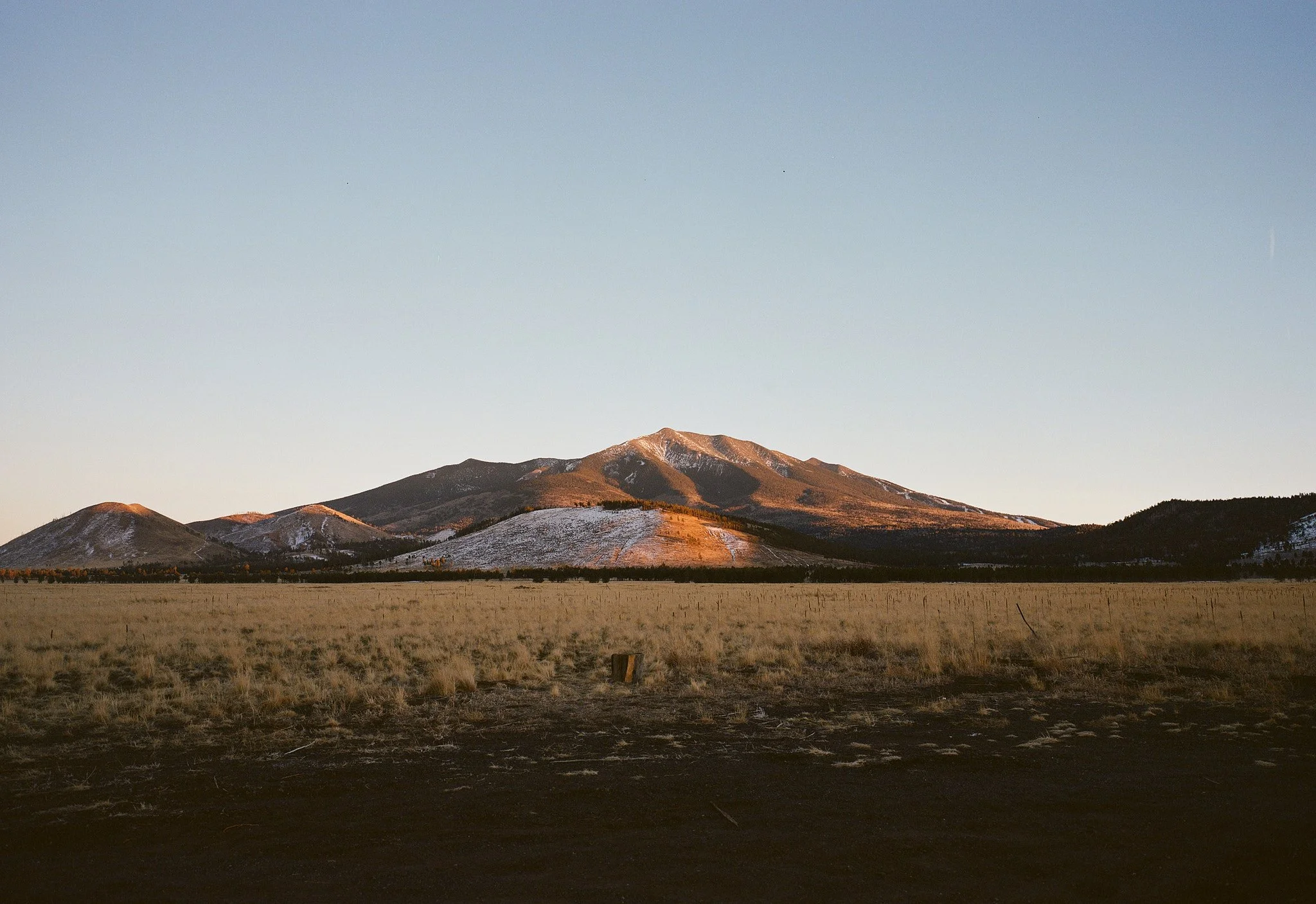 A mountain range with a snowy peak at sunrise or sunset, surrounded by grassy plains and a clear blue sky.