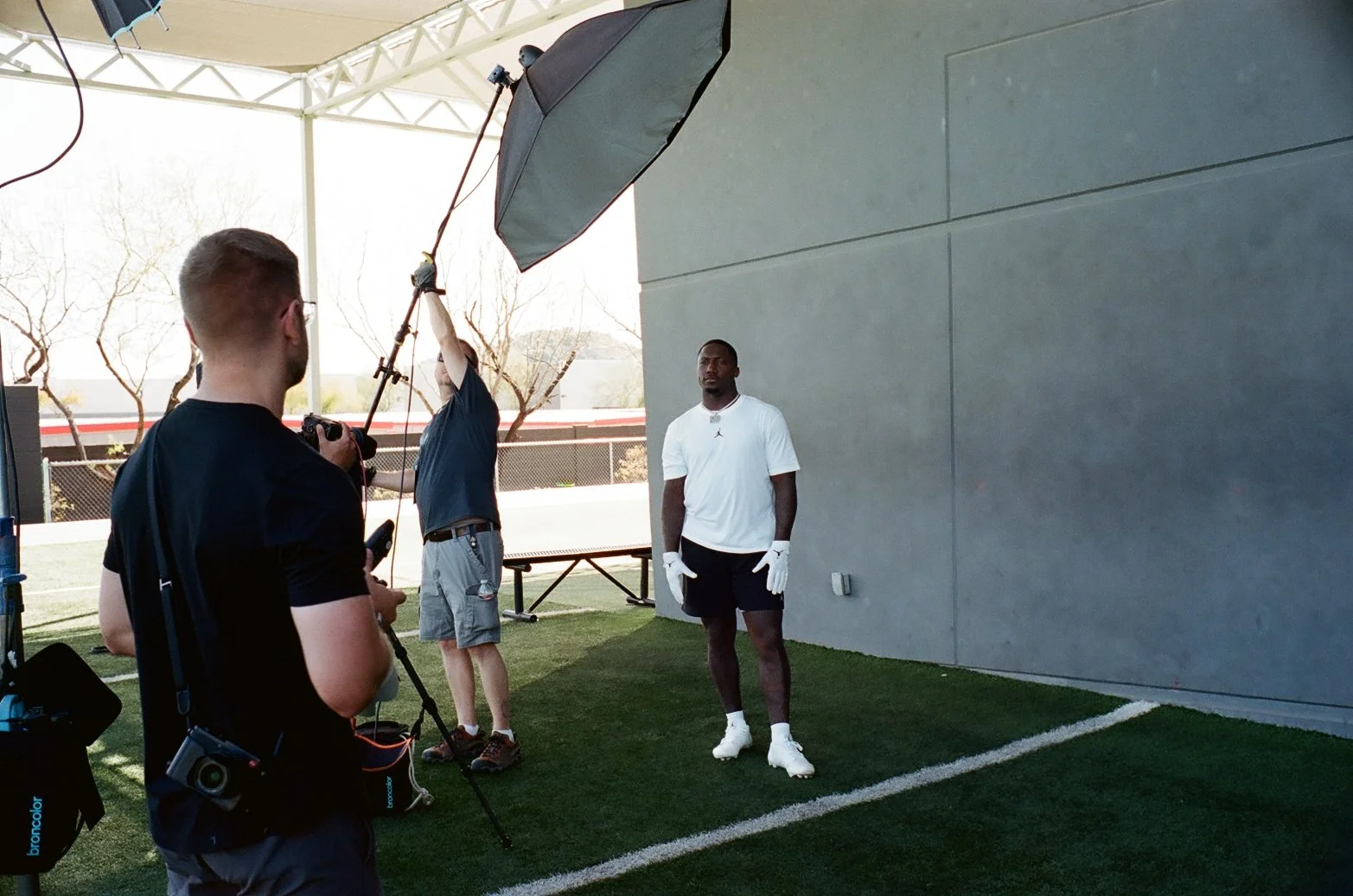 A photoshoot on a green turf field, with a male athlete in white shirt and shorts standing against a gray wall, and a team of crew members setting up lighting and camera equipment.