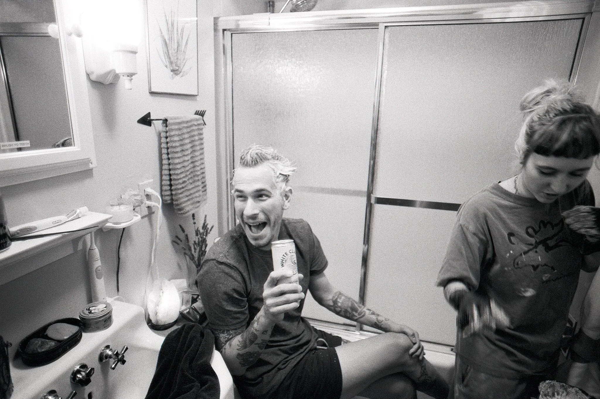 A group of people in a bathroom, with one person sitting on a counter holding a can and smiling, while another person is mixing something in a bowl.