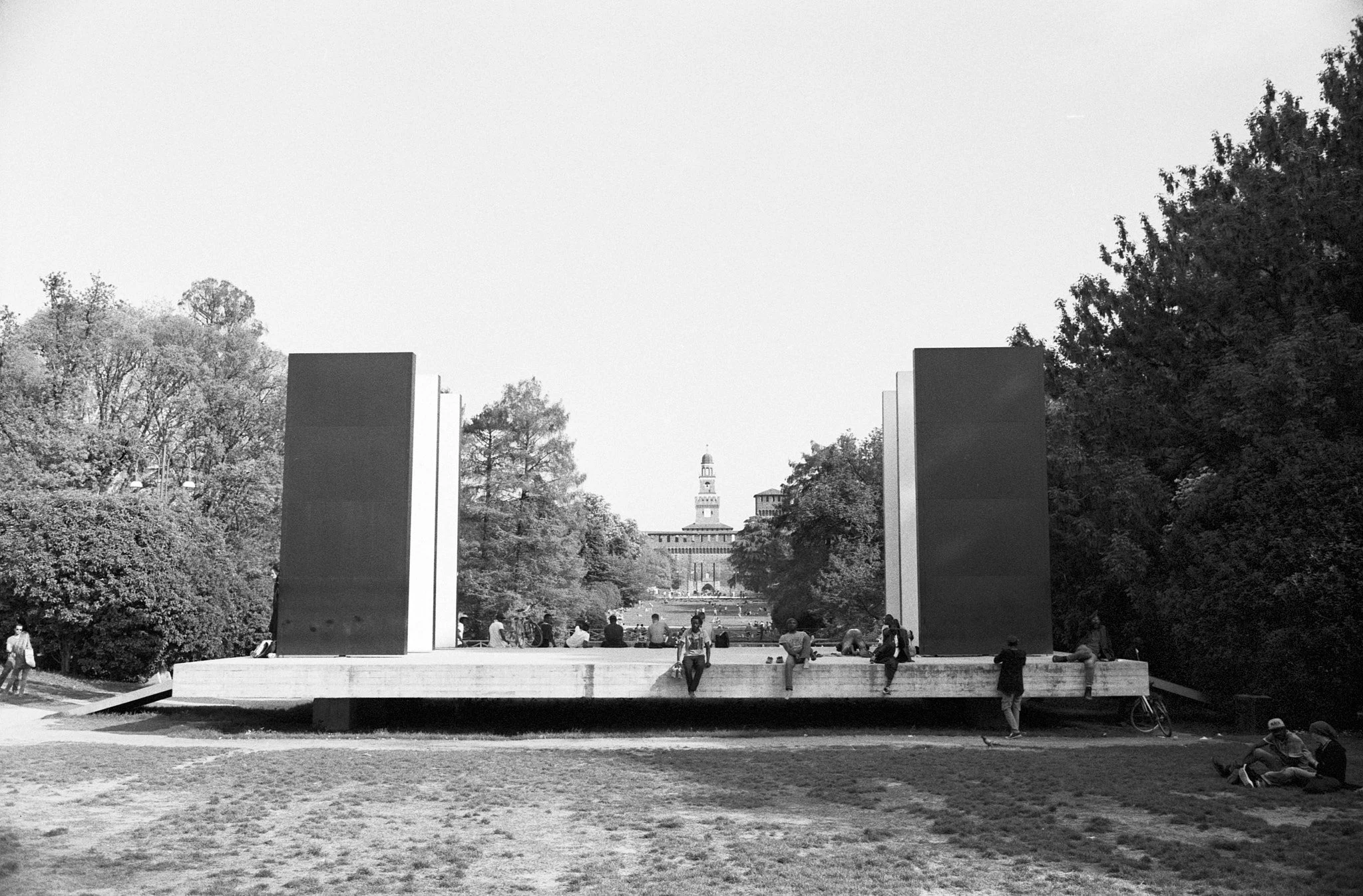 People sitting and relaxing on a modern outdoor stage or pavilion in a park with trees and a historical building with a tower in the background, black and white photo.