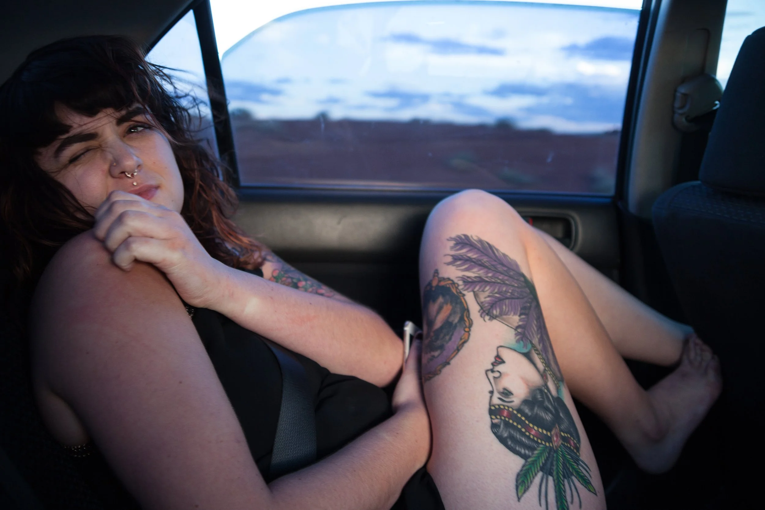 A woman with tattoos sitting in the back seat of a car, looking at the camera, with an outdoor landscape visible through the window.