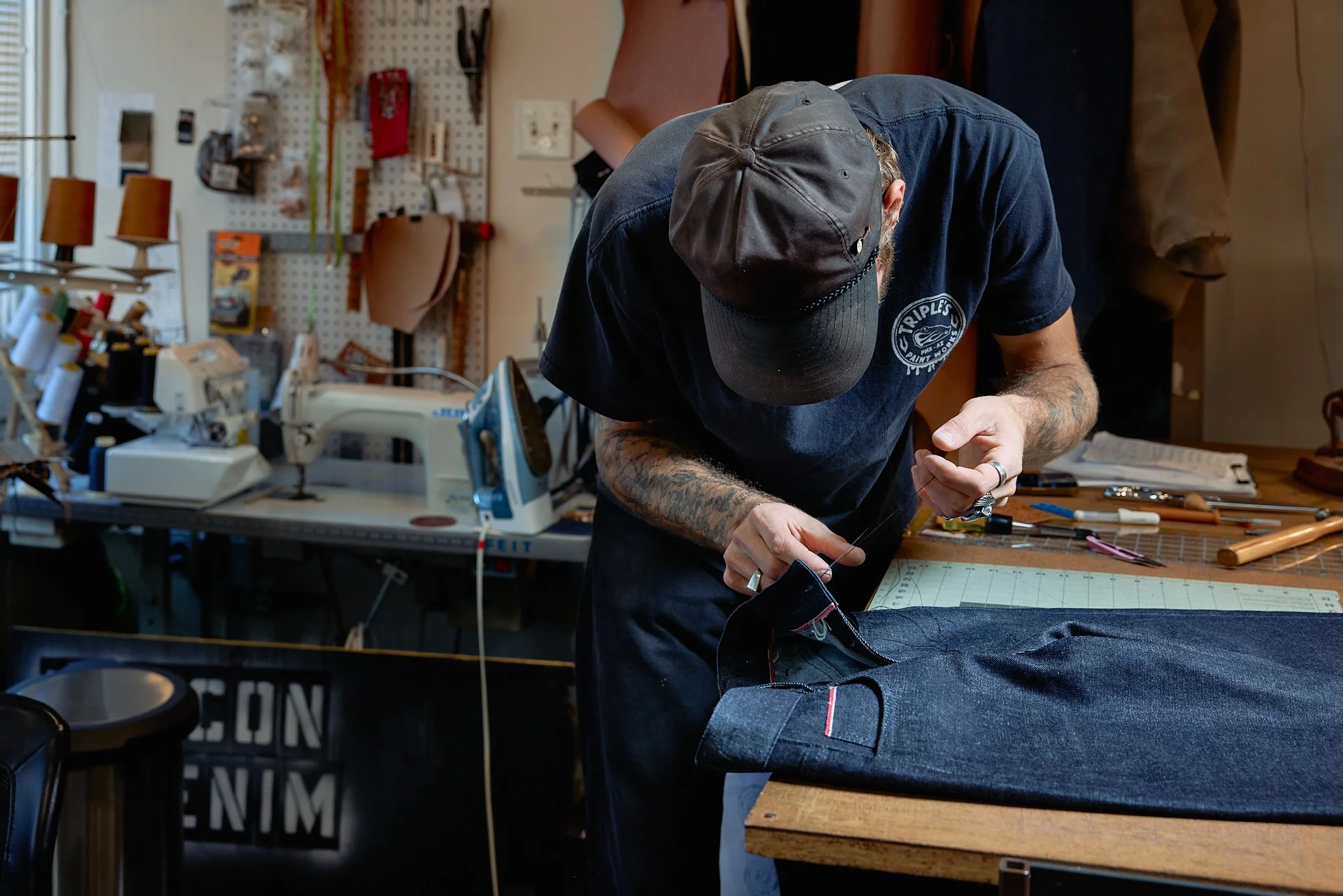 A person with tattoos and wearing a baseball cap is sewing fabric on a work table in a workshop with sewing tools, an iron, and a pegboard in the background.