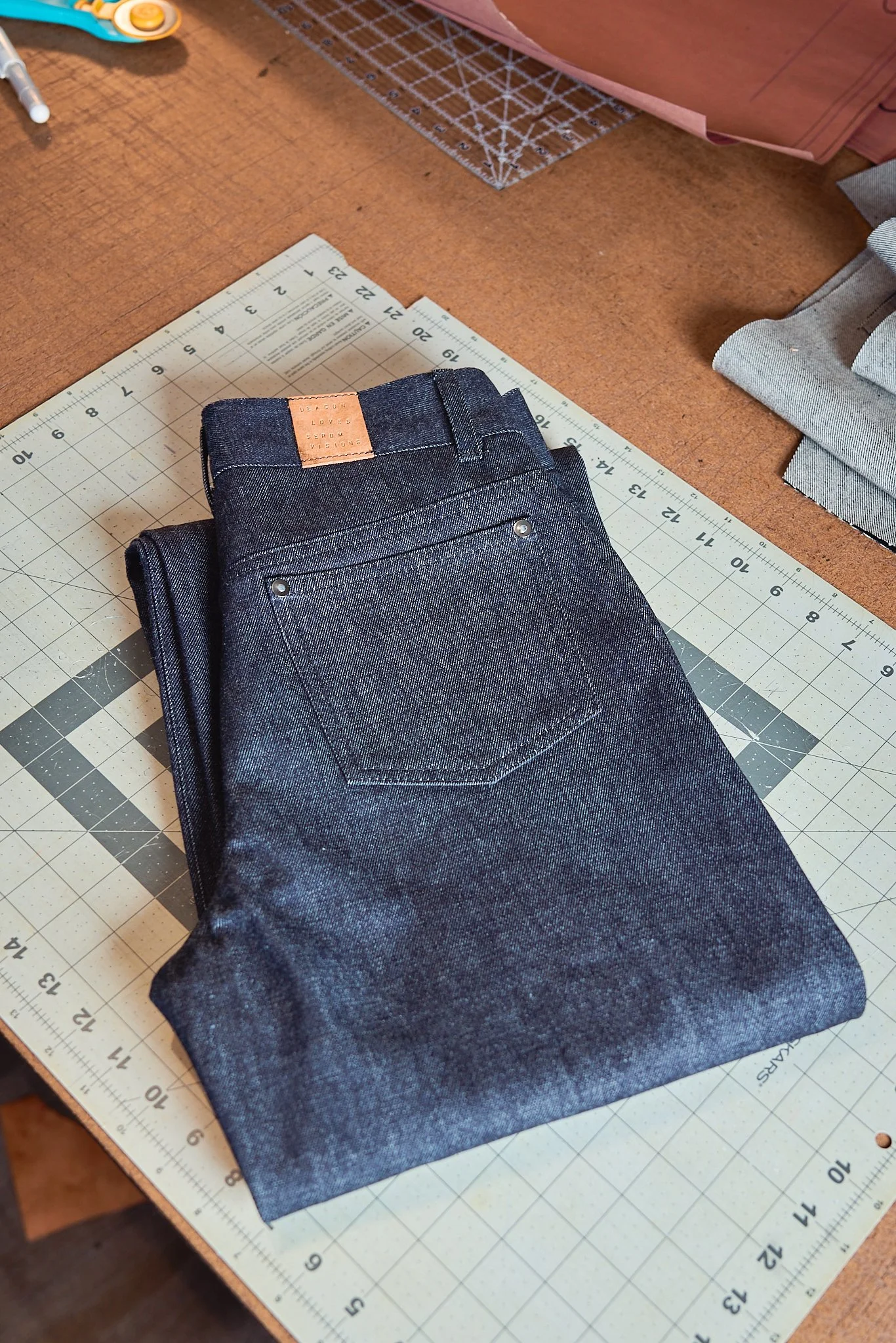Folded pair of dark blue jeans with a tan leather patch on the waistband, placed on a cutting mat on a worktable.