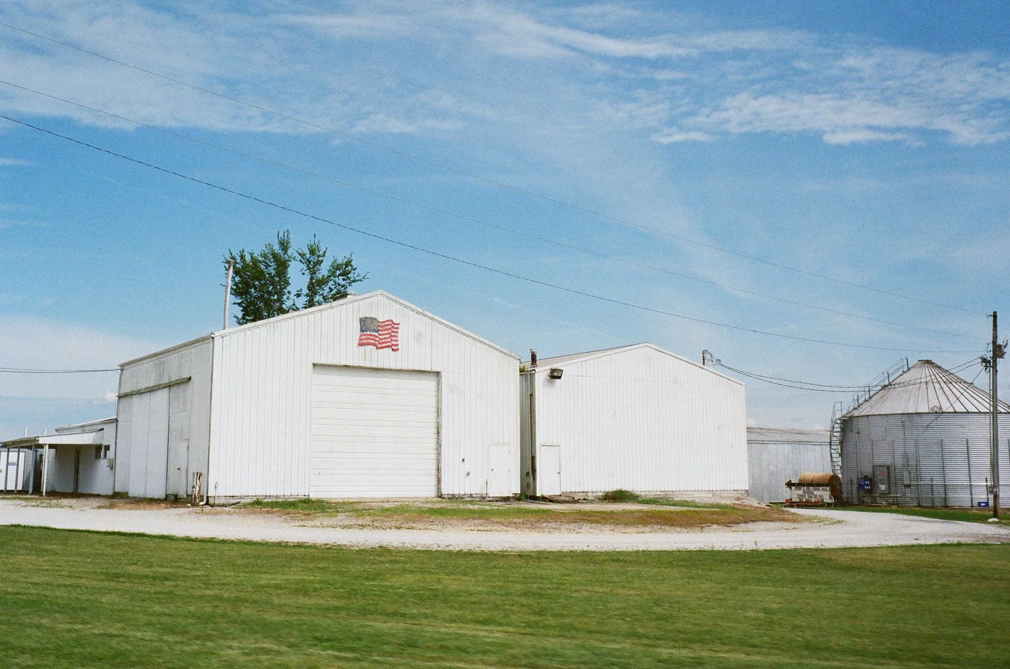 A white industrial building with an American flag painted on the front, set against a blue sky with some clouds, surrounded by grass and gravel, with silos on the right.