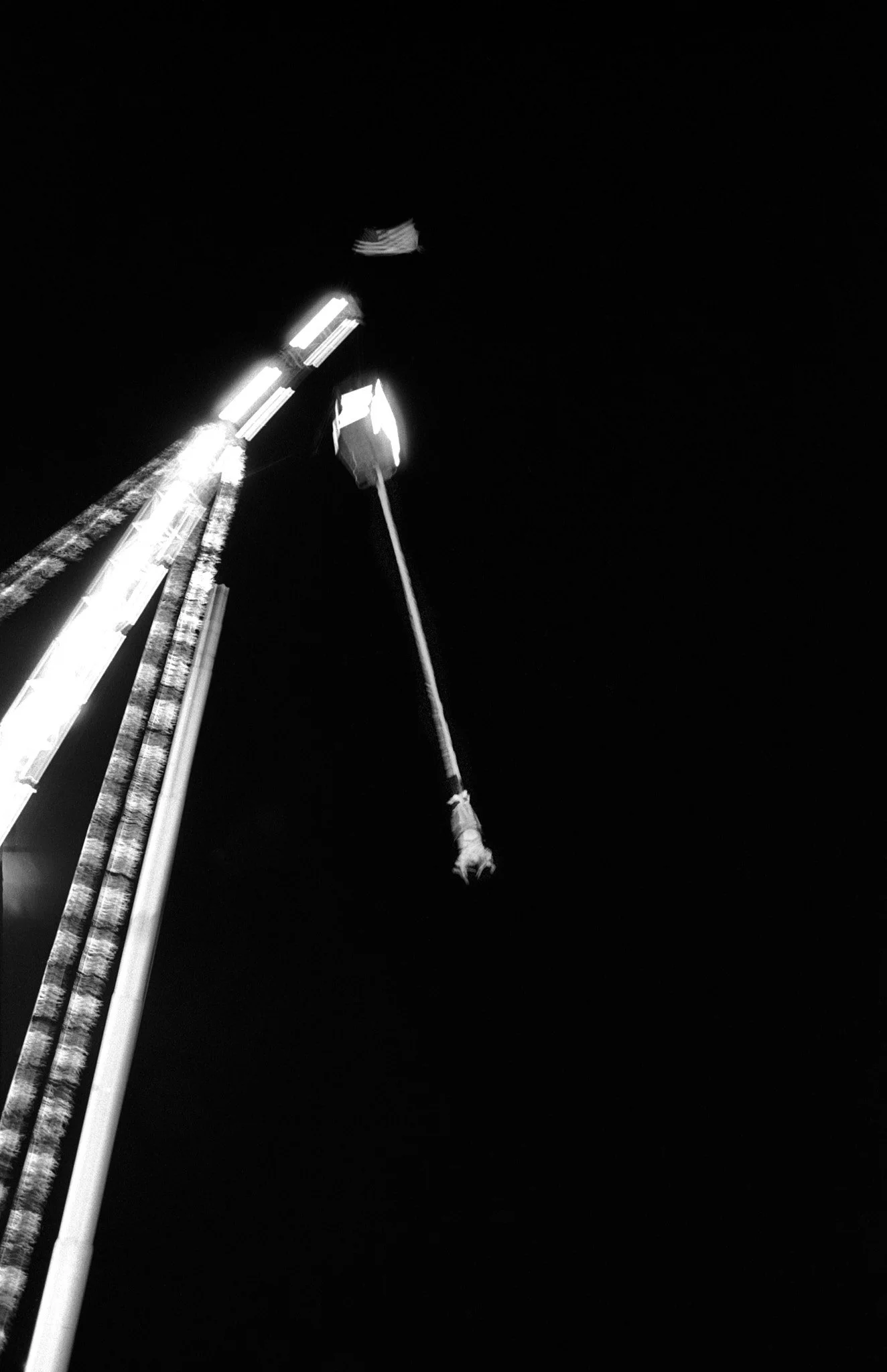 A black and white photo of a tall amusement park ride at night with bright lights, featuring a swinging cage attached to a tall tower.