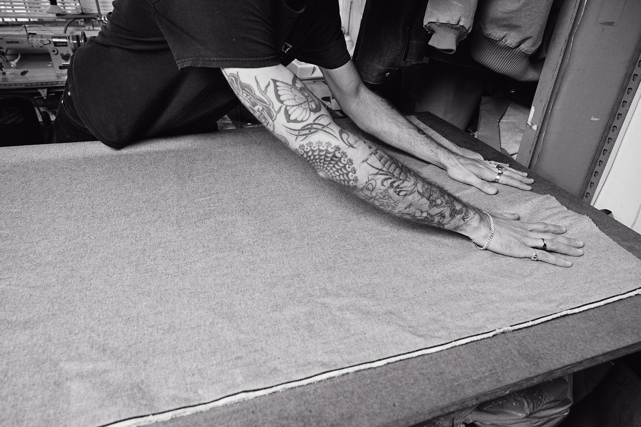 A person with tattoos on their arms is working on fabric on a table in a workshop.
