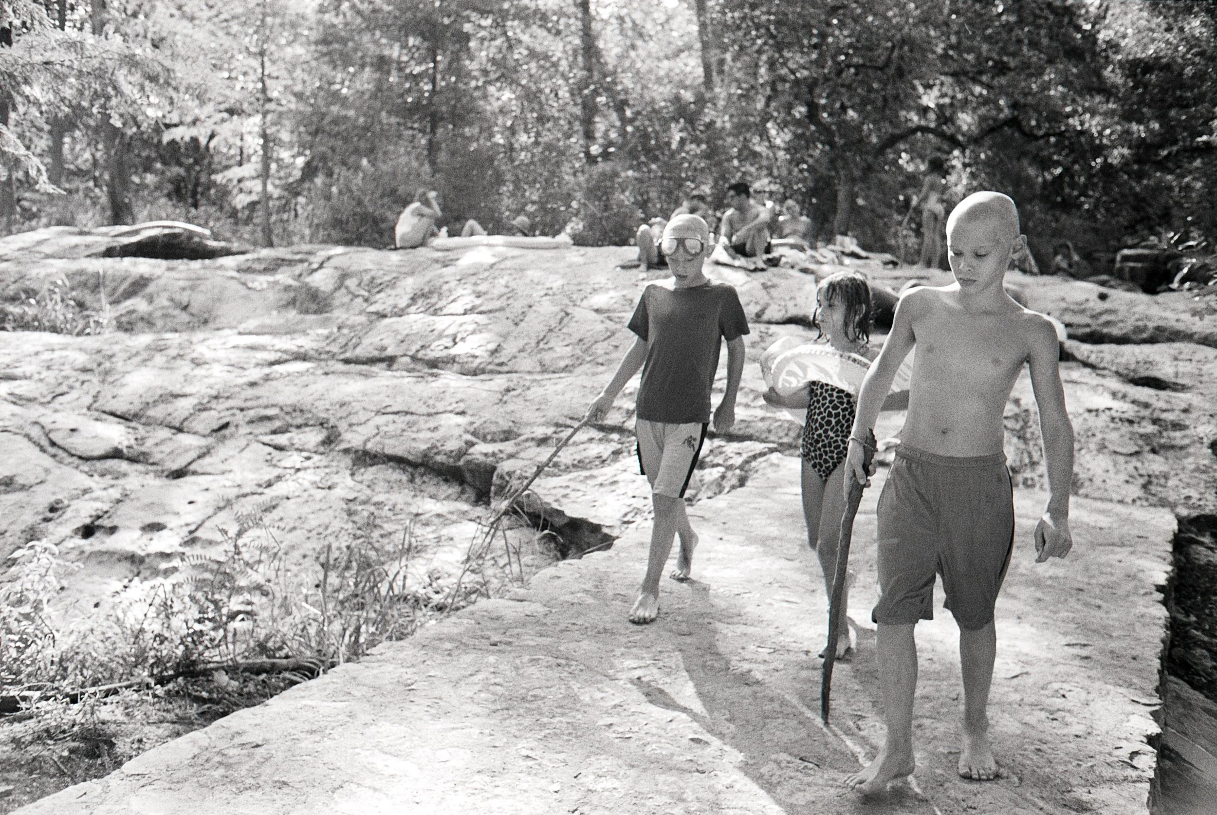 Children walking barefoot on a rocky path near a body of water, some holding sticks. Trees and people sitting on rocks in the background.