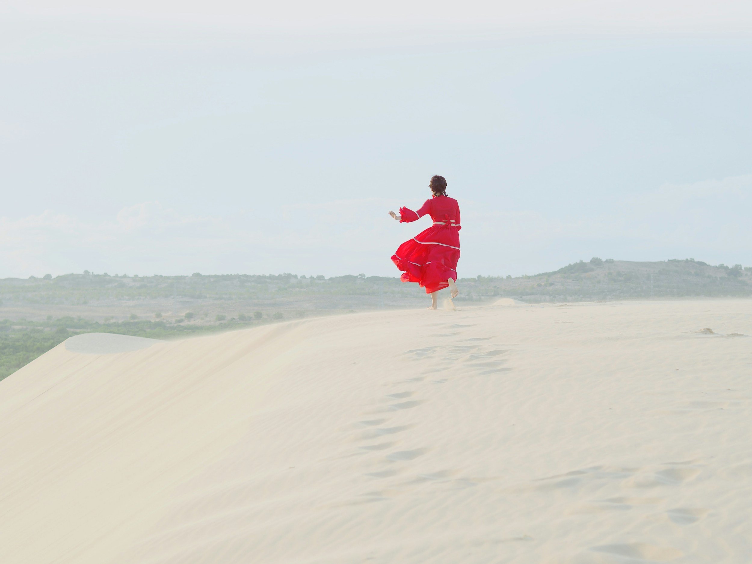 A person in a long, flowing red dress running on a sandy beach under an overcast sky.