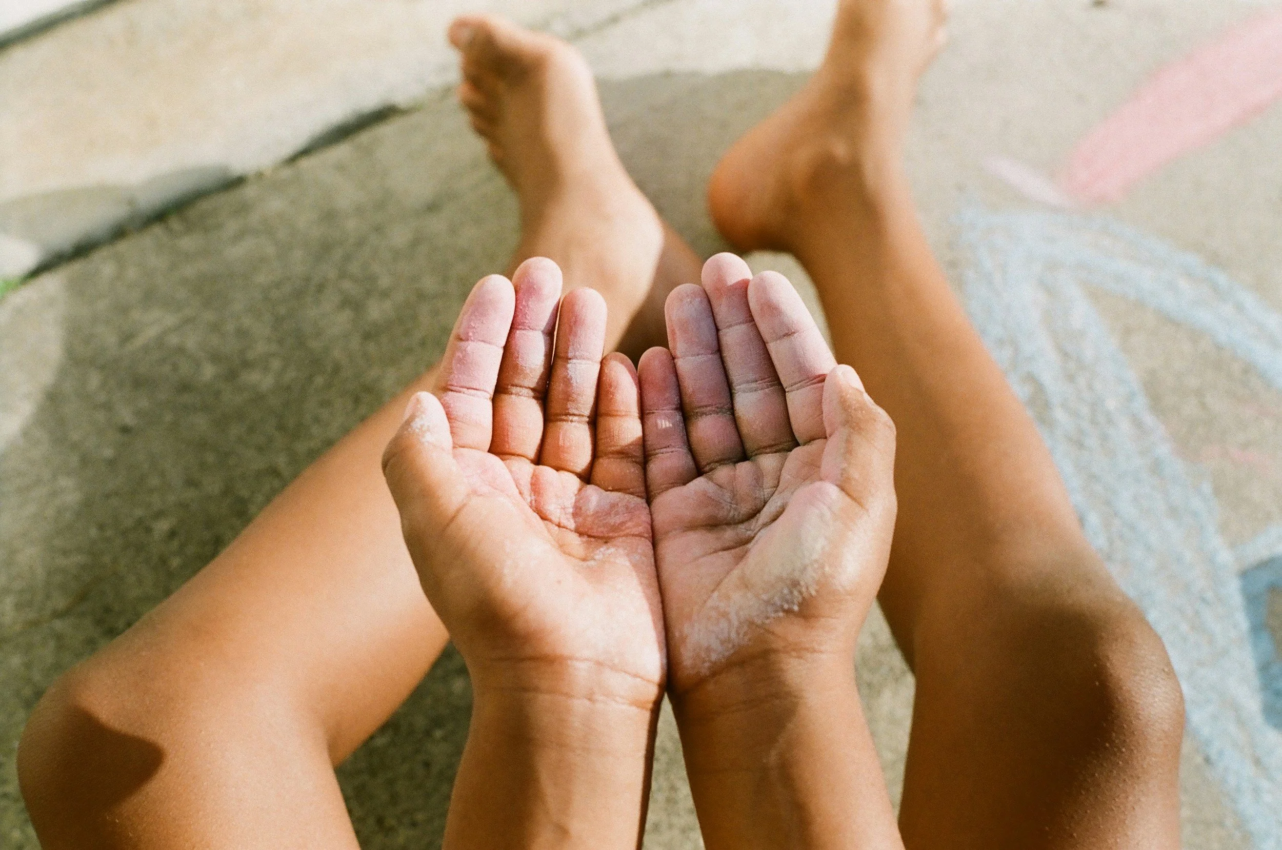 A person with dark skin and dirt-covered hands holds an open pair of hands, also with dirt, in front of their legs on a beige pavement.