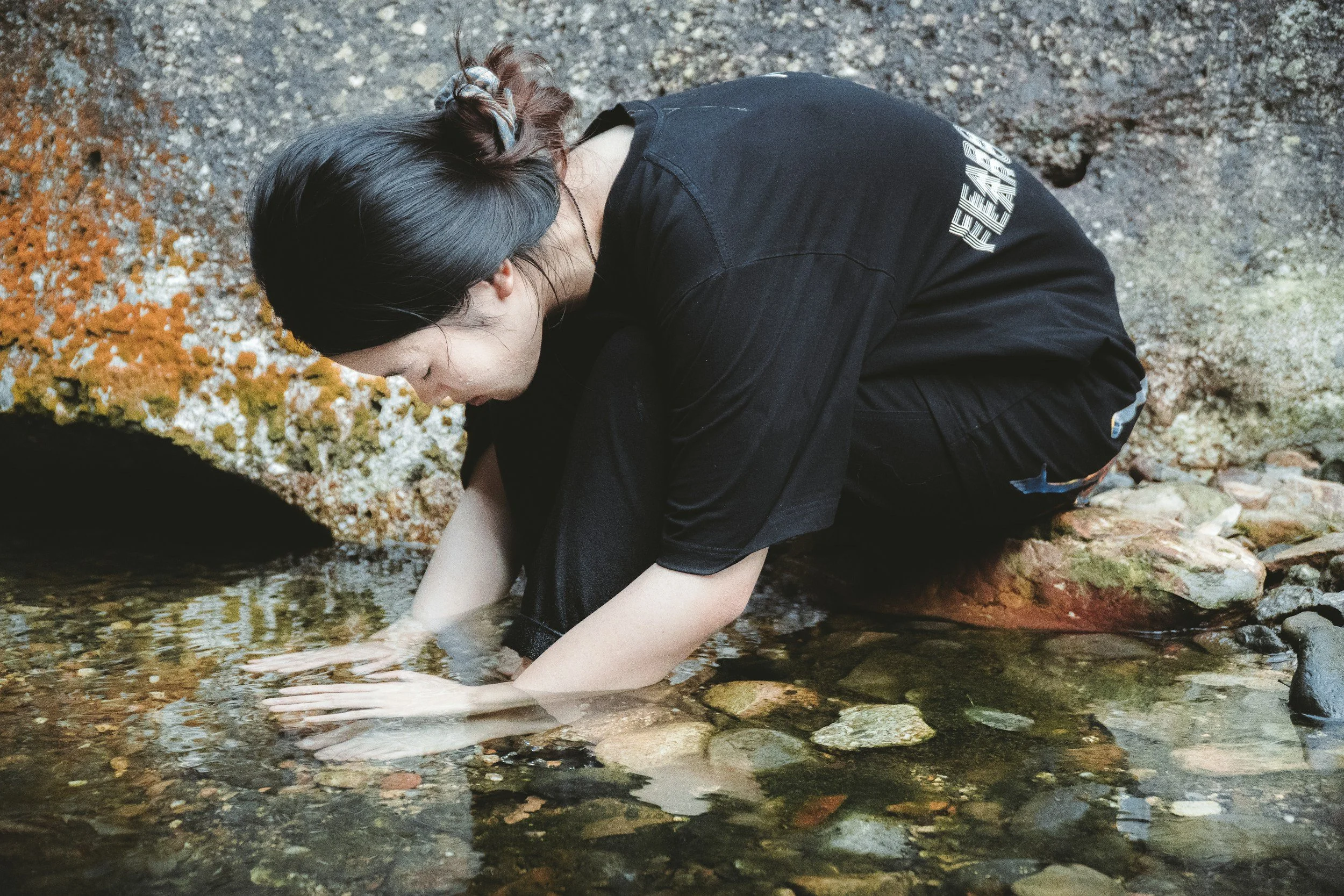 A woman washing her hands in a small creek with rocks and autumn foliage in the background.