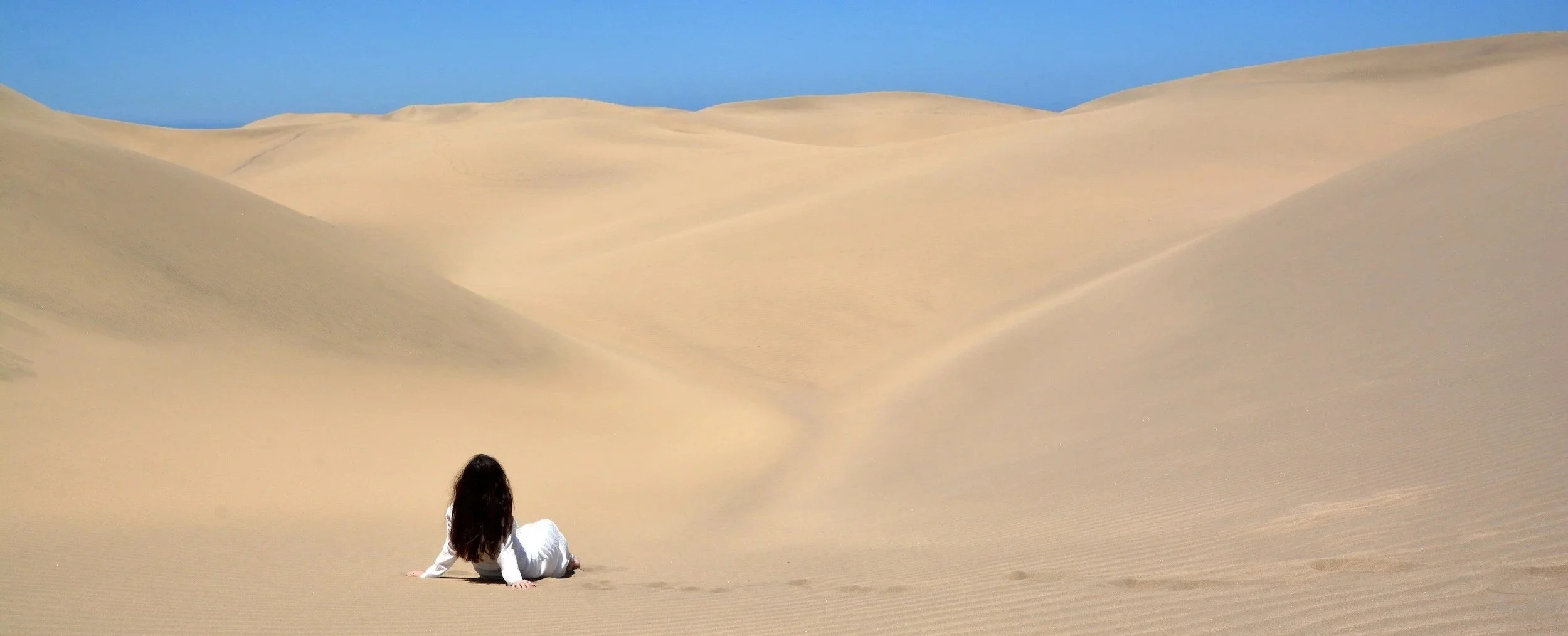 Person sitting on sand dunes in a desert, facing away from the camera, with a clear blue sky above.