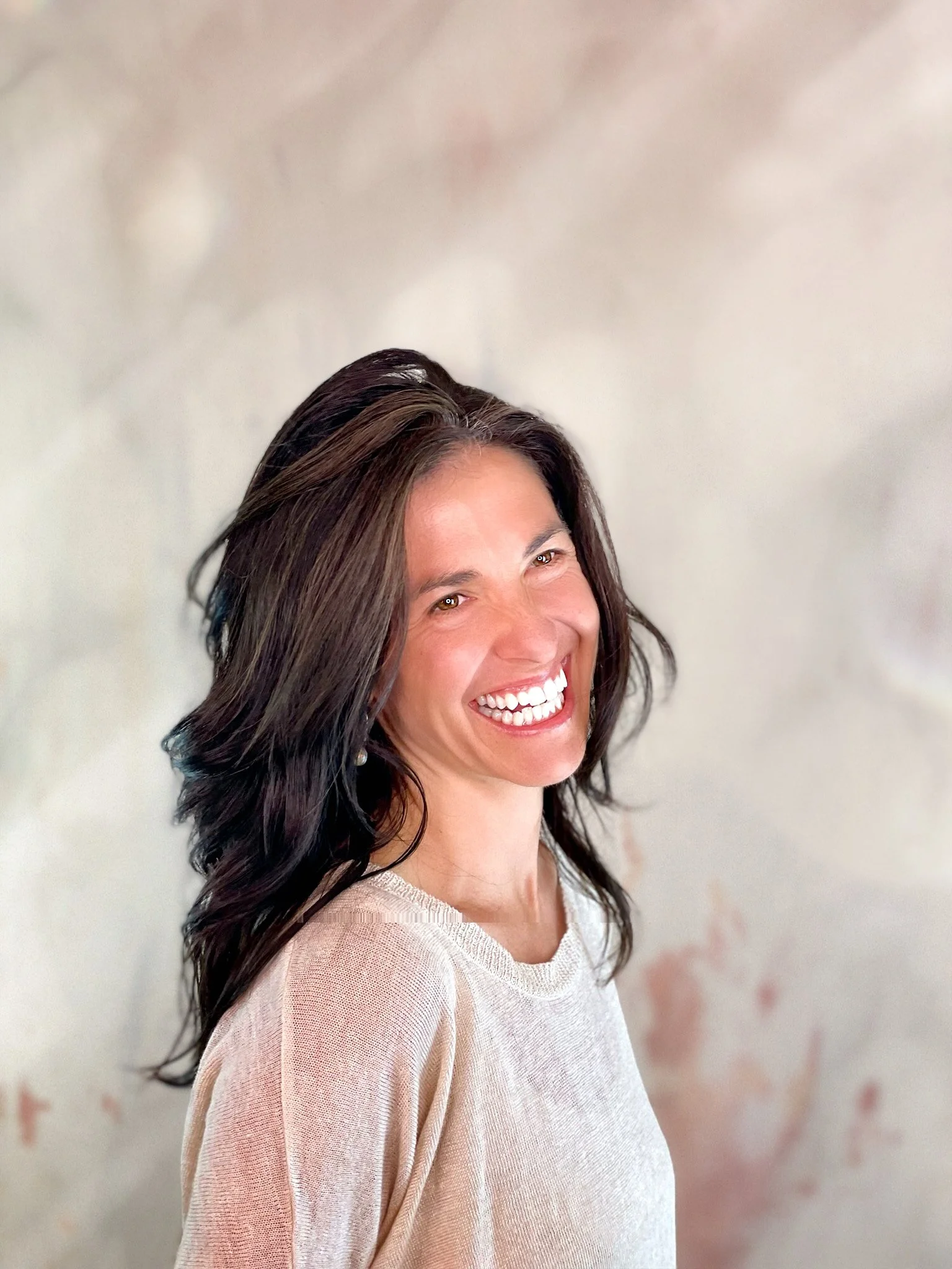Smiling woman with dark wavy hair, wearing a beige sweater, standing against a soft, blurred background.
