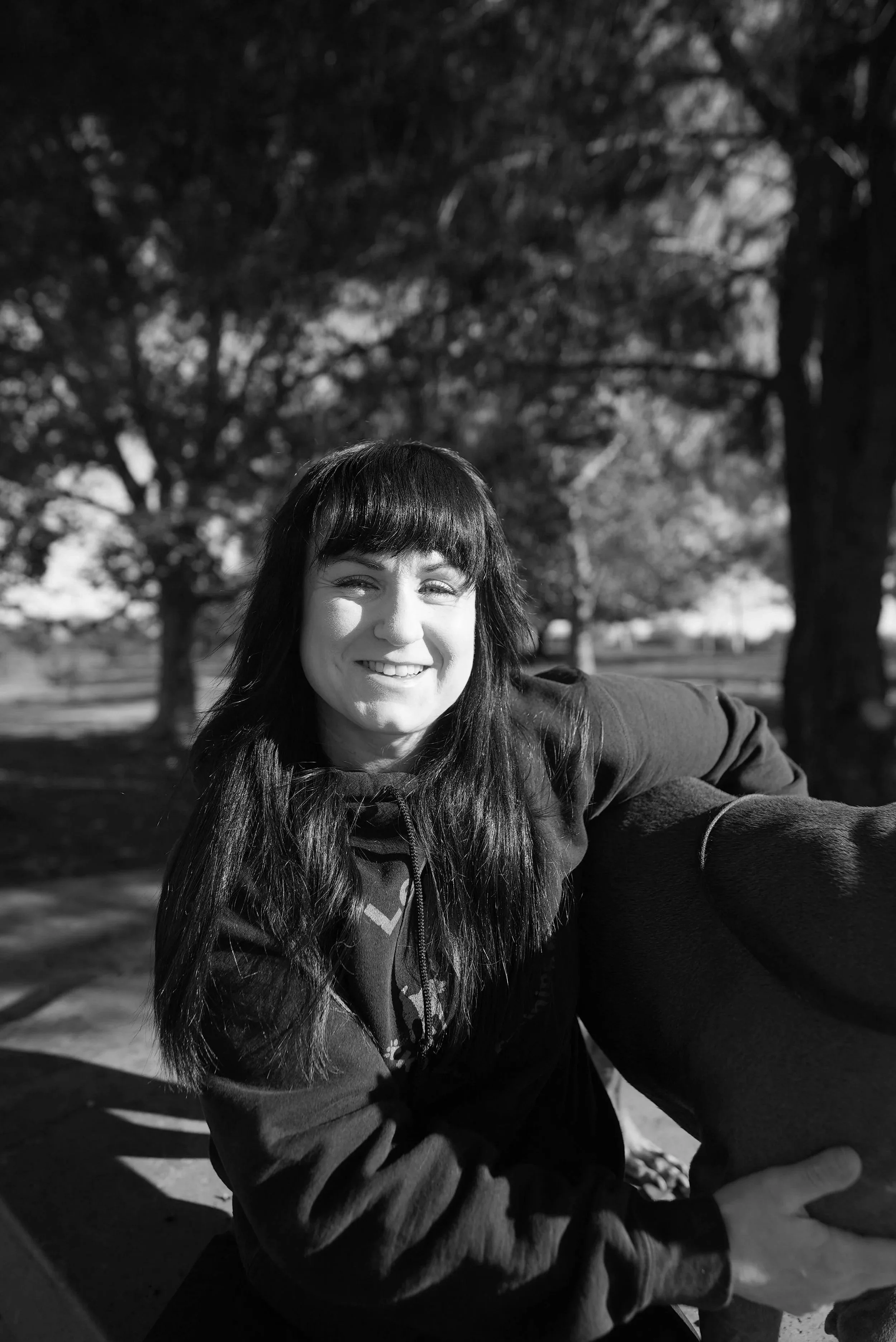 A woman with long dark hair smiling outdoors in a park with trees in the background, taking a selfie.