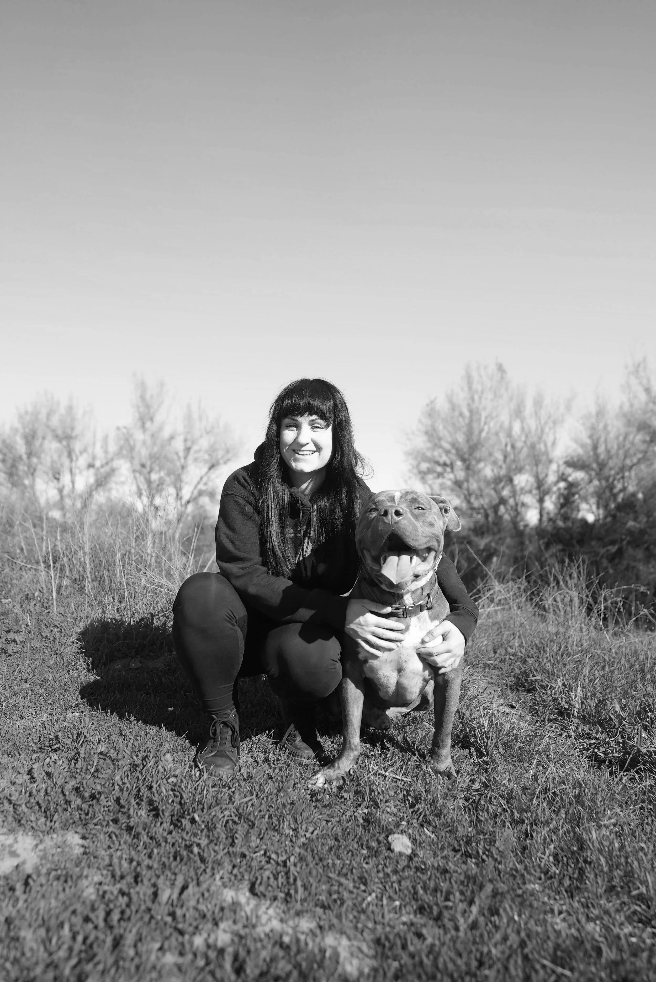 A young woman with dark hair and bangs crouches beside a happy, excited dog outdoors, with trees and a clear sky in the background.