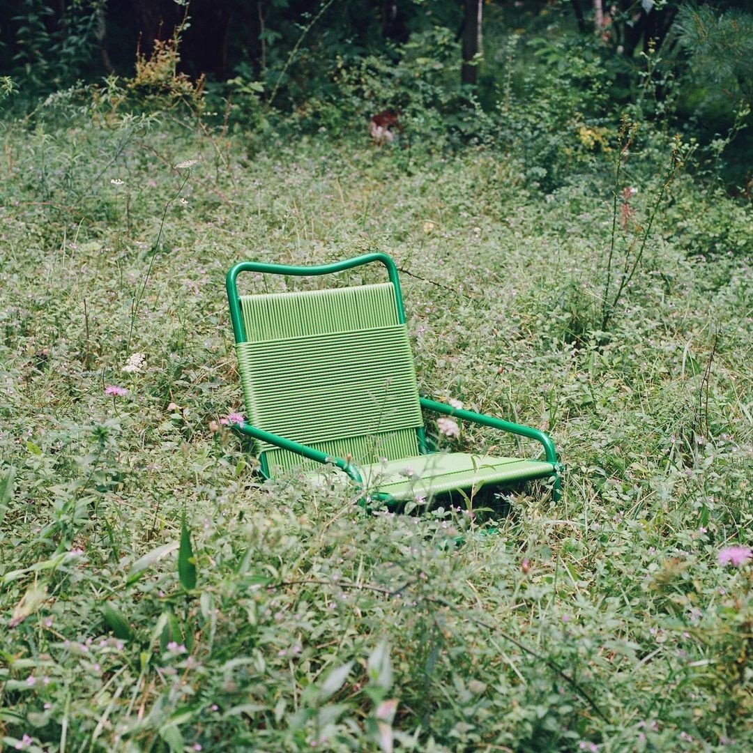 A green outdoor chair with a metal frame and woven seat, left in overgrown grass and wildflowers, with trees in the background.