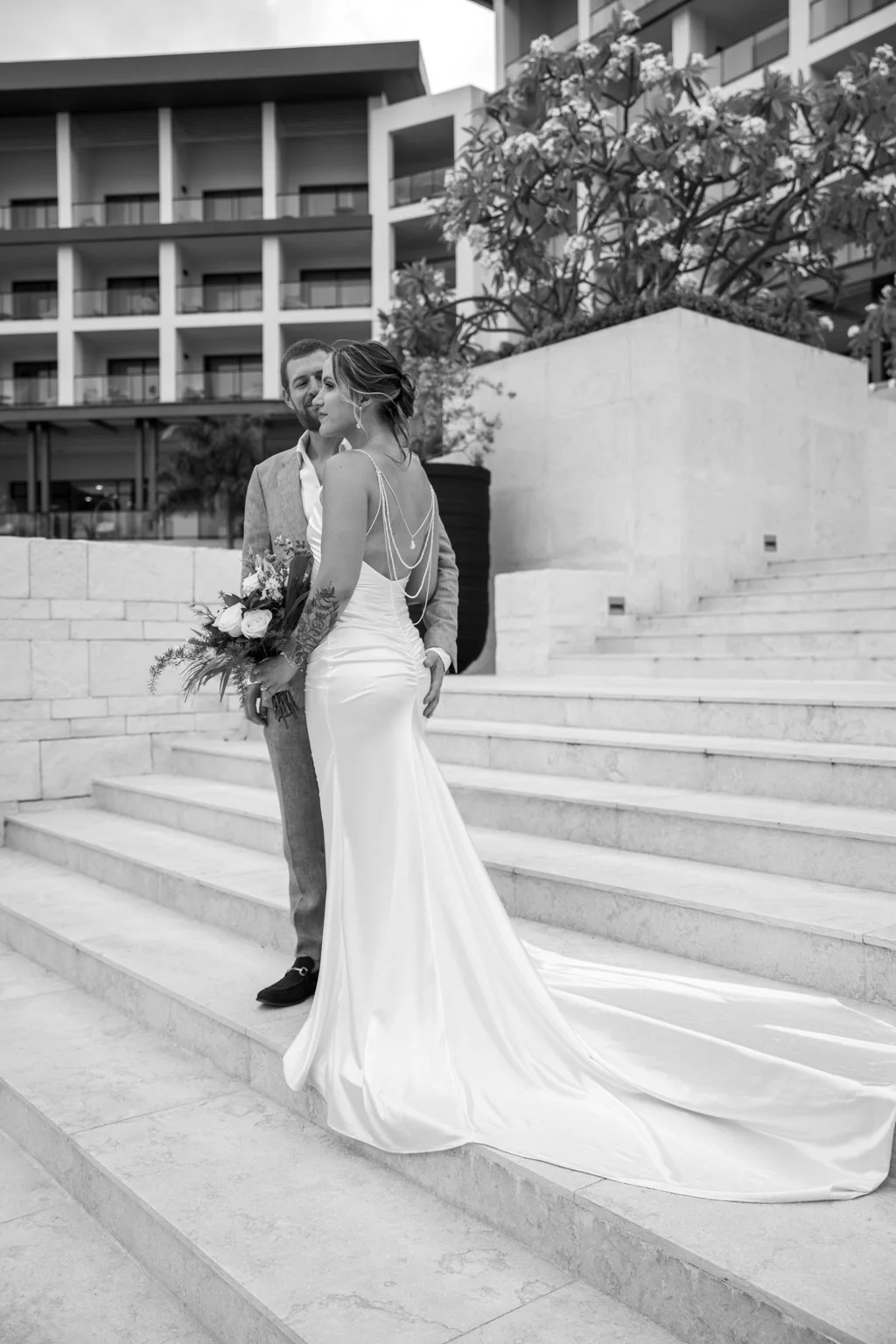 A bride and groom stand on stairs outside a modern building, with the bride holding a bouquet of flowers. The bride wears a long, elegant wavy white wedding dress with thin straps and decorative chains on her back. The groom wears a light-colored suit with a white shirt and black shoes. The scene is in black and white.