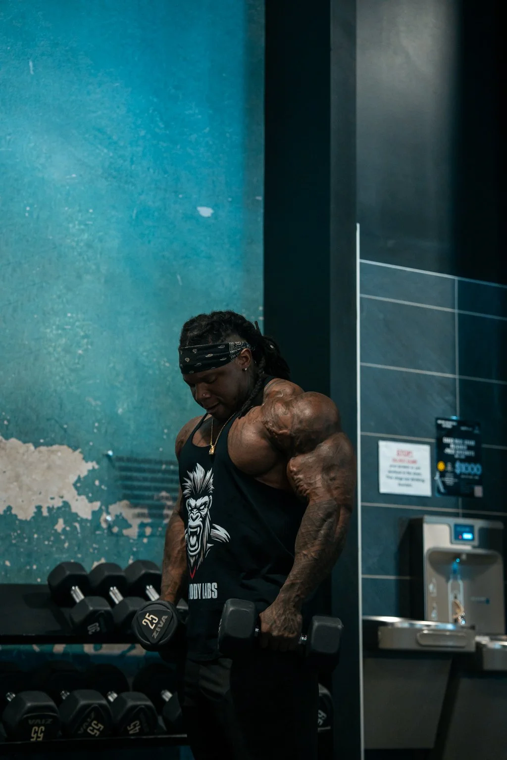 Male fitness model lifting a dumbbell in a gym setting, wearing a black tank top and bandana with teal textured walls and gym equipment behind him.