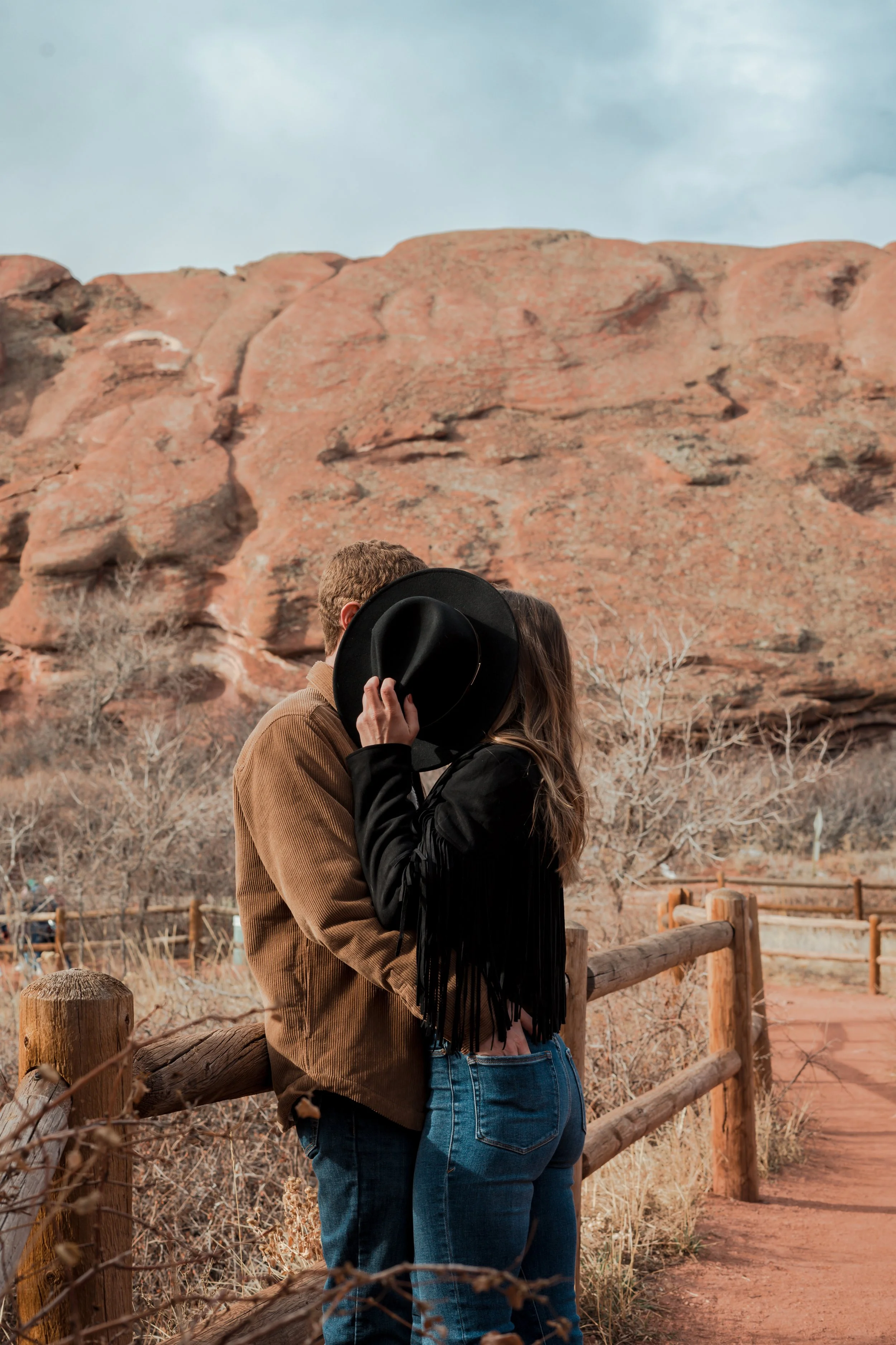 A couple kissing outdoors near a rocky, desert-like landscape with a dirt pathway and wooden fence.