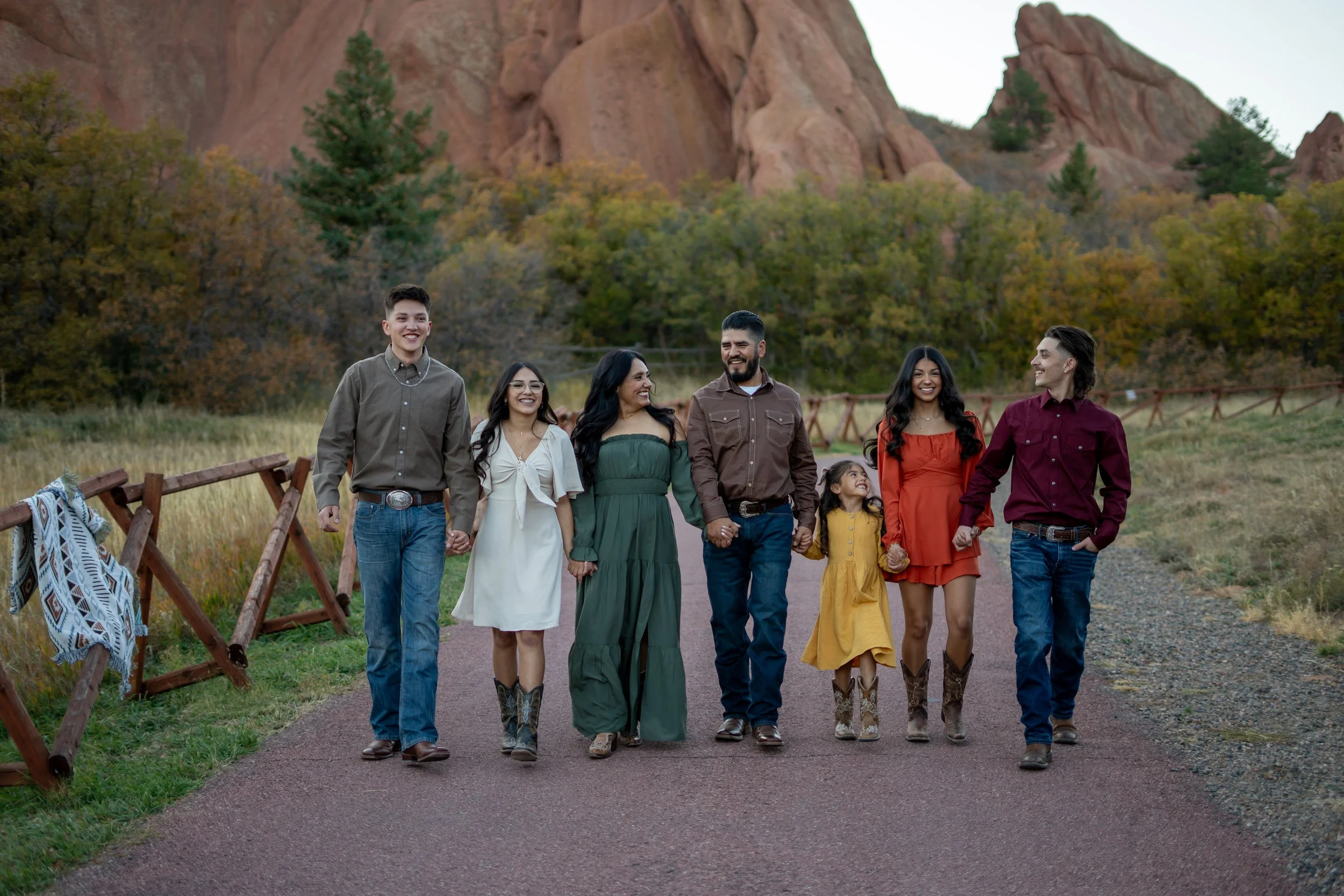 A multi-ethnic family of seven walking hand in hand outdoors on a dirt path, with red rock formations and trees in the background. The family is dressed in semi-casual clothing, and they are smiling and enjoying each other's company.
