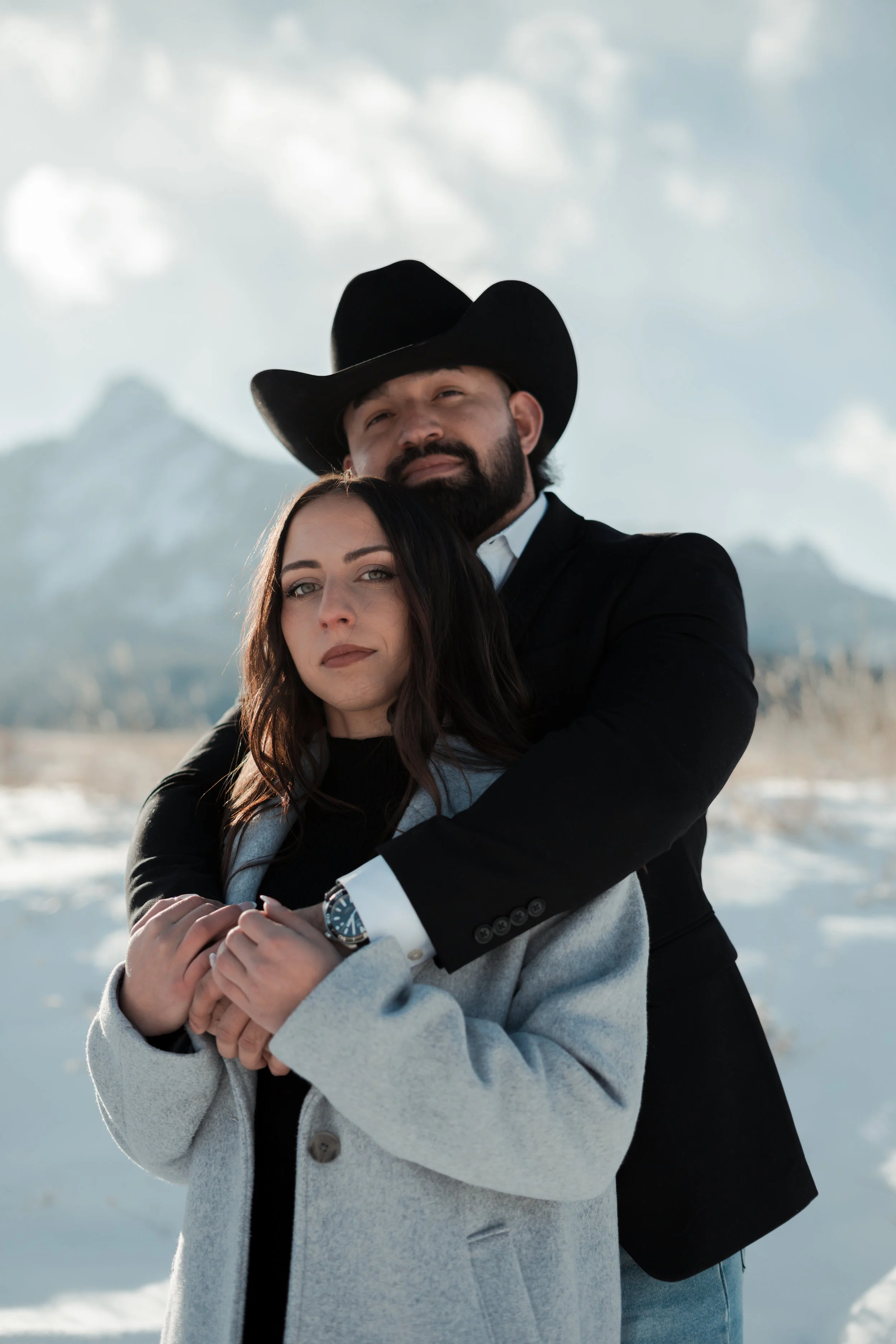 A man wearing a cowboy hat and a woman in a gray coat standing in a snowy landscape with mountains in the background, embracing each other. Proposal. Engagement shoot
