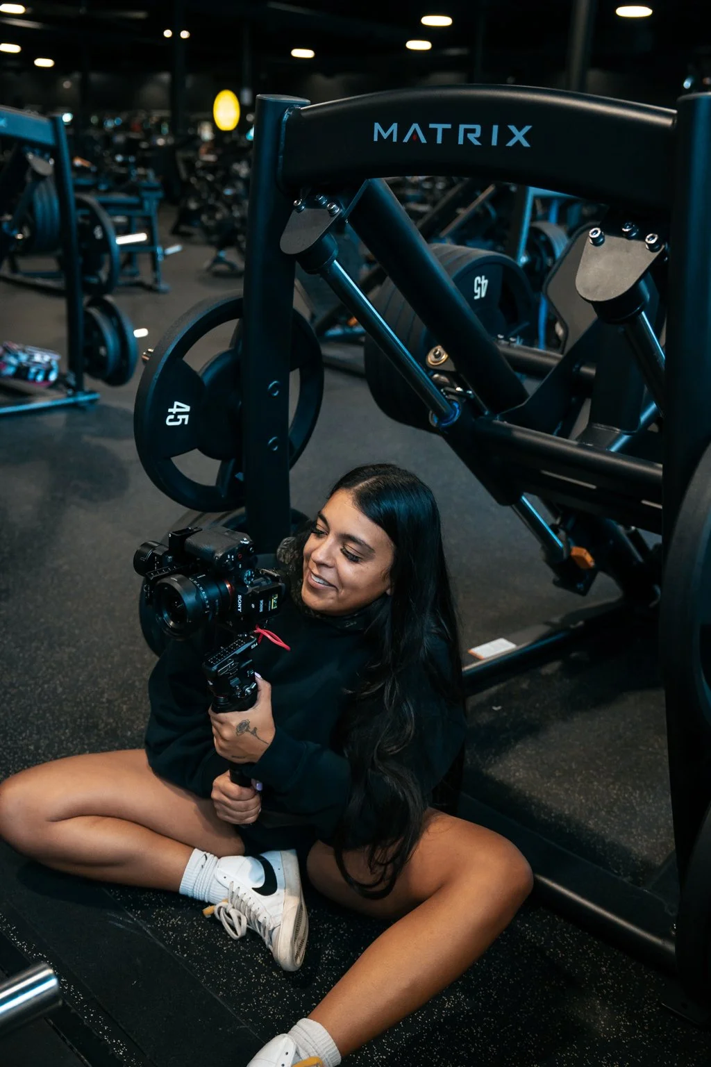 A woman sitting on the gym floor holding a camera, with a large weight machine overhead and gym equipment in the background.