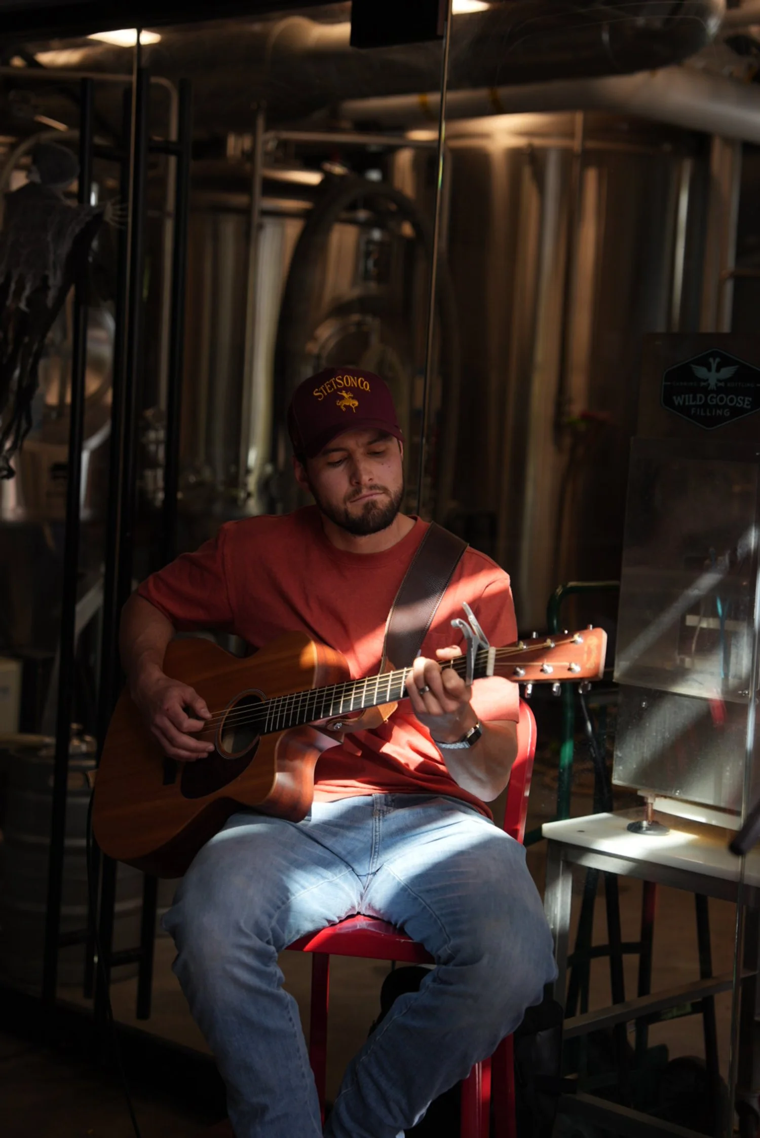 A man wearing a maroon cap and red t-shirt playing an acoustic guitar in a dimly lit setting.