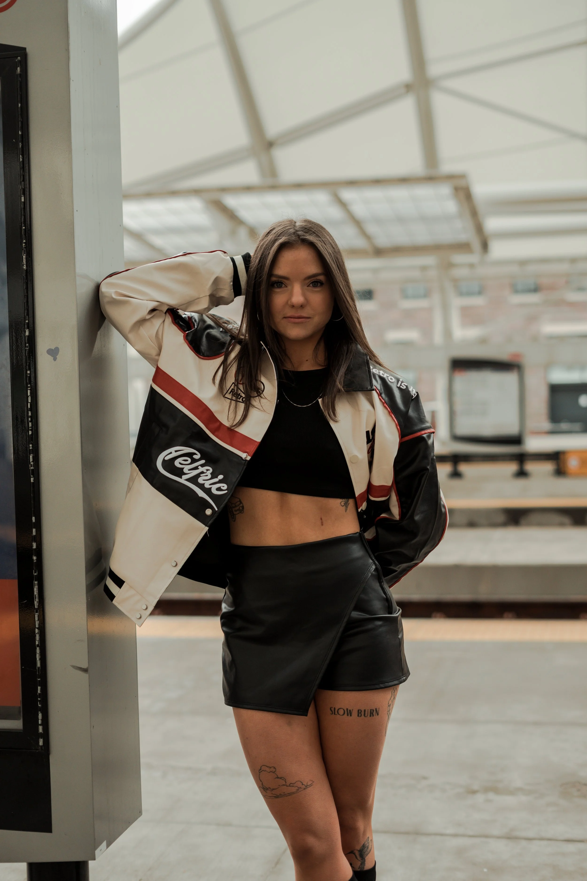 Young woman wearing a black crop top, a varsity jacket, and a black leather skirt, standing at a train station.