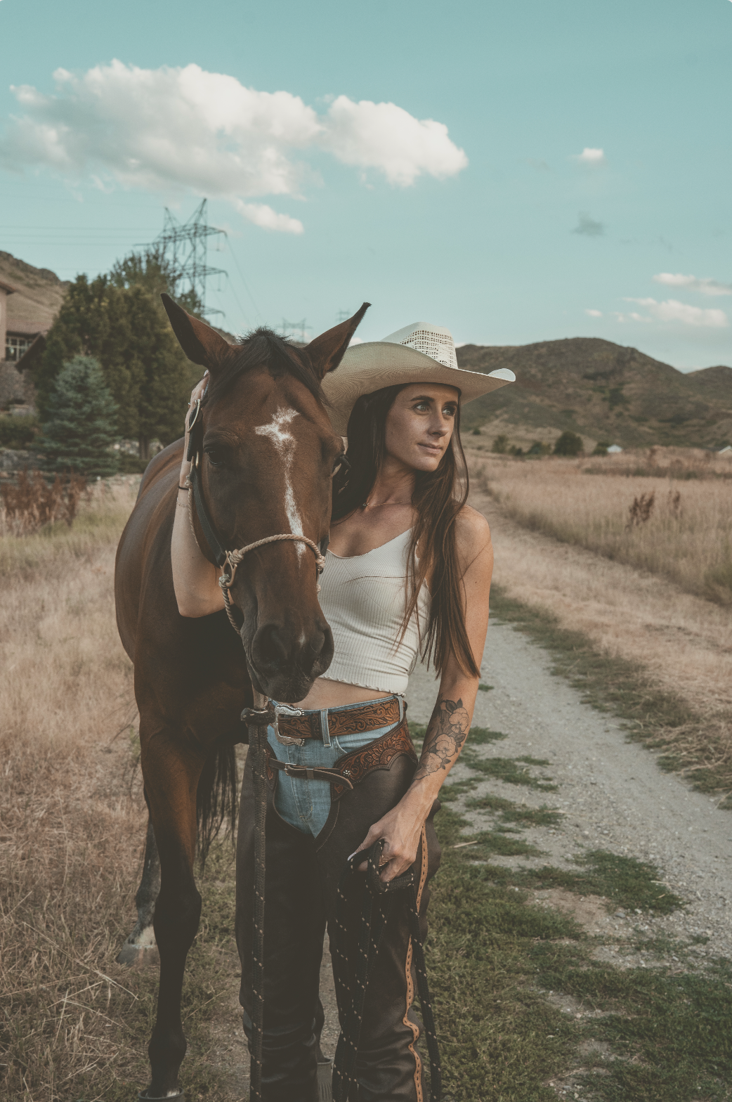 A woman in a cowboy hat standing beside a brown horse on a rural dirt road in a valley with hills in the background.