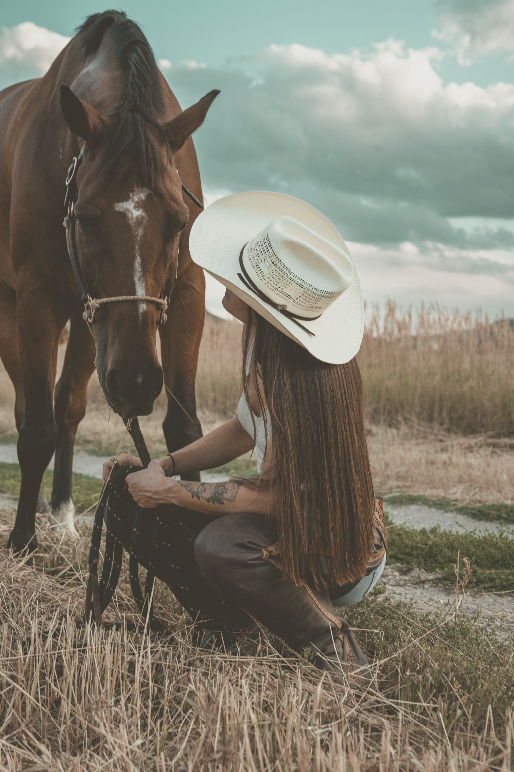 A woman with long brown hair wearing a white hat and white shirt kneels on the grass, holding the bridle of a brown horse with a white star on its forehead, in a field with tall grass and a cloudy sky in the background.