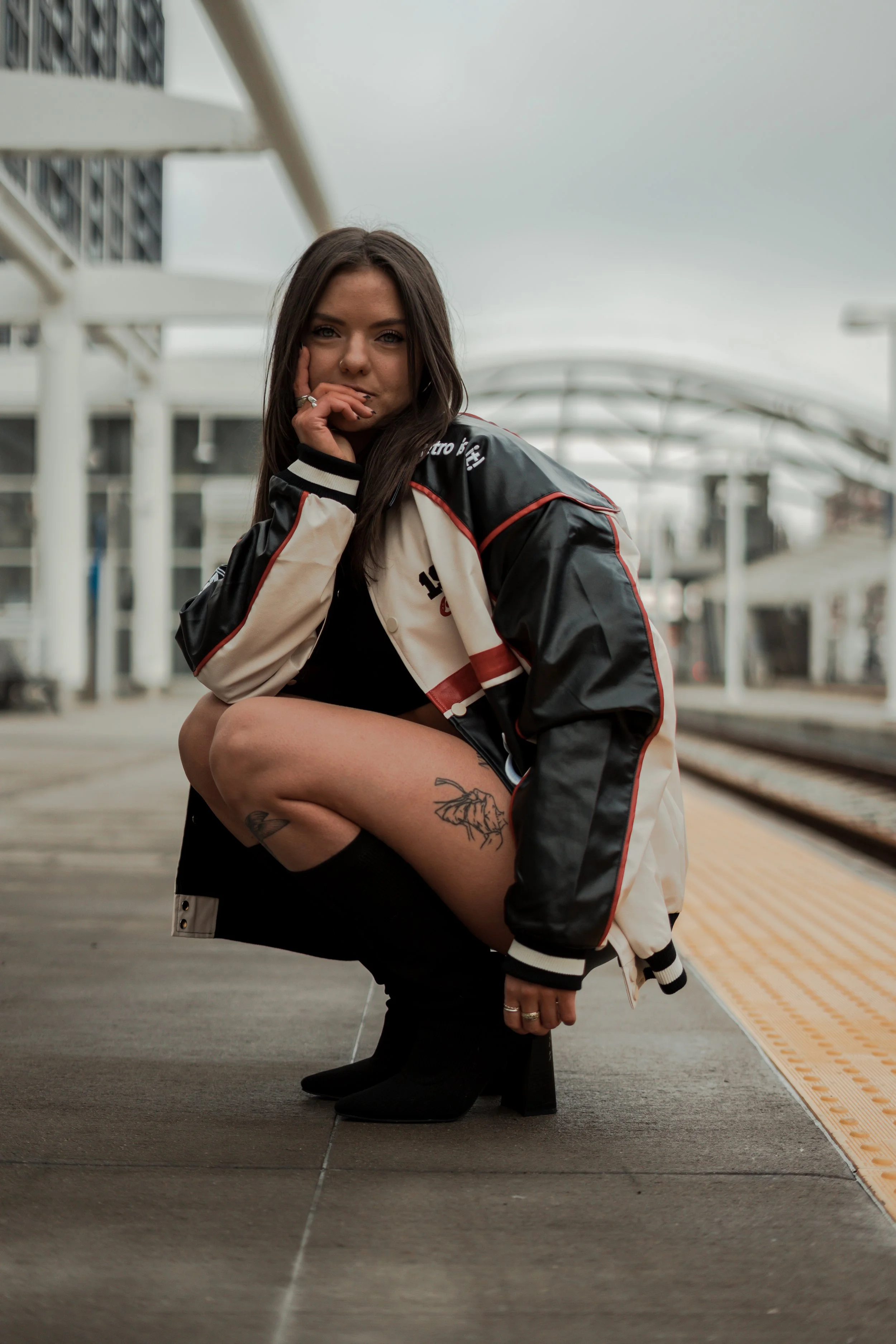 A young woman wearing a black dress, black high-heeled boots, and a varsity jacket crouches on a train platform, looking at the camera with a hand near her face. She has tattoos on her thigh and a piercing on her nose. The background shows train trac