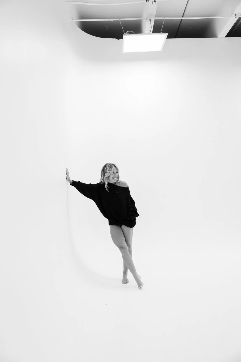 A woman in a black sweater and light-colored pants smiling and posing against a plain white background in a photography studio with overhead lighting.
