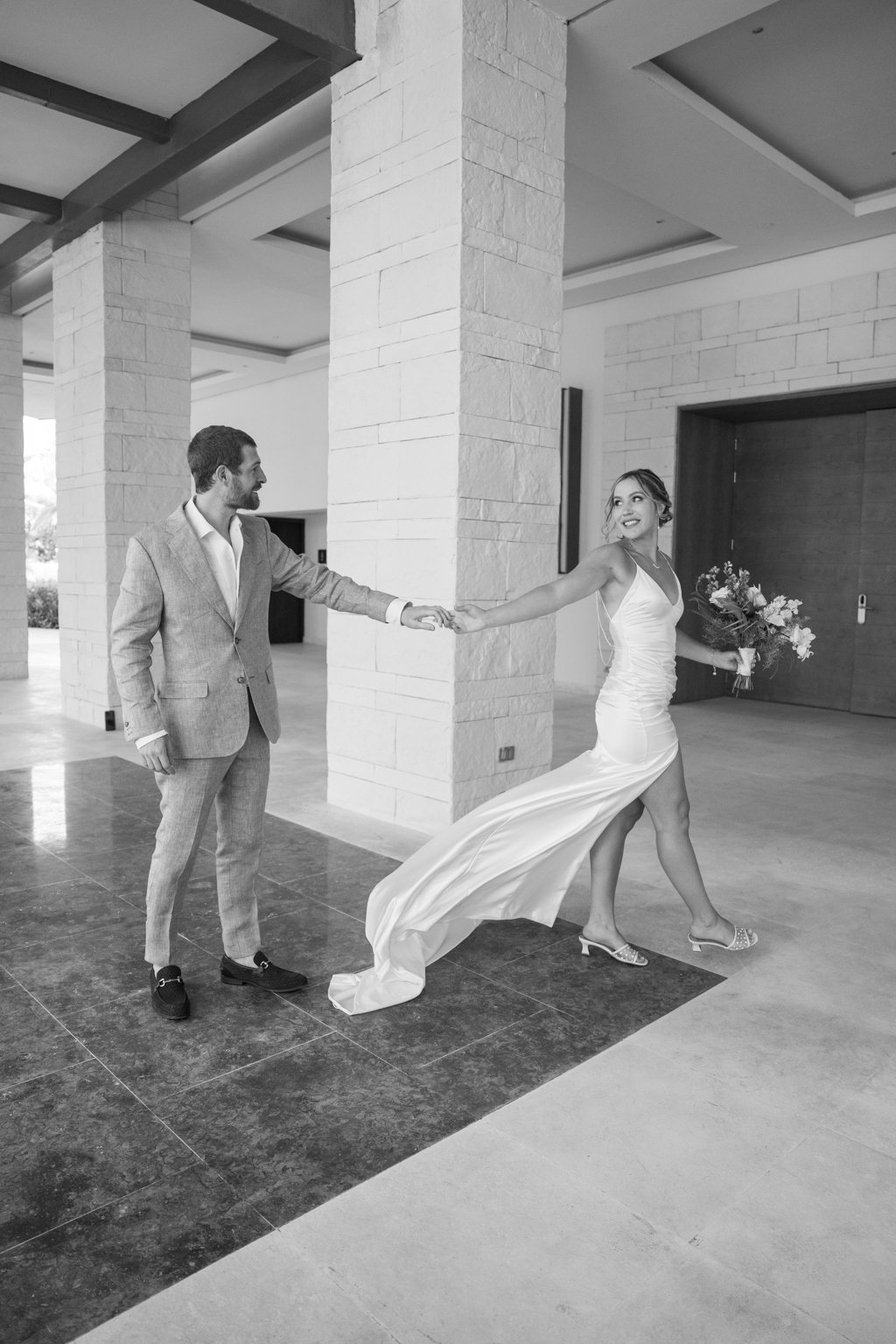 A bride in a wedding dress holding a bouquet of flowers is playfully pulling a groom in a suit by the hand inside a modern building with stone walls and an open space.