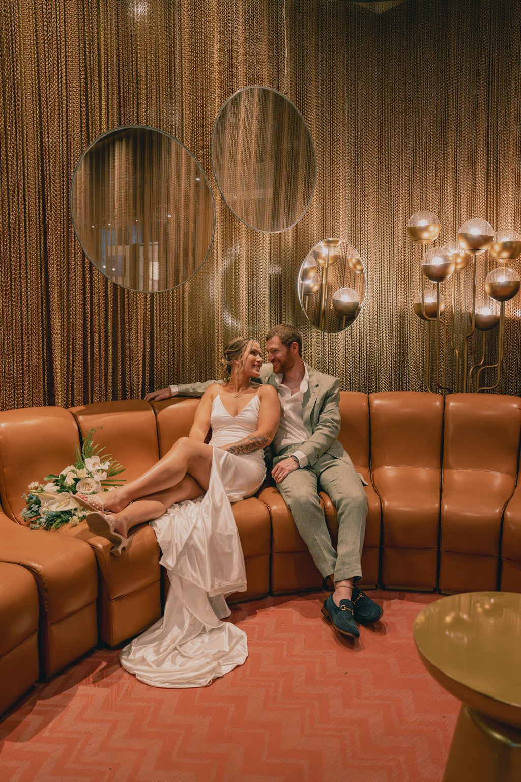 A couple in wedding attire sitting on a brown leather couch in a stylish, warmly lit room with gold tones, modern wall mirrors, and decorative lighting fixtures.