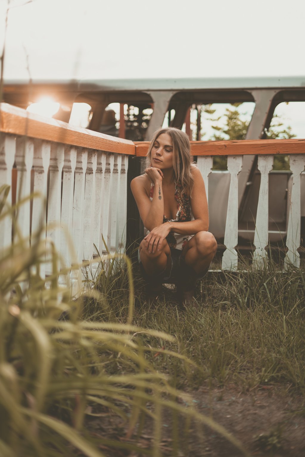 A woman crouches on grass near a white wooden fence, with a vintage trailer in the background during sunset.