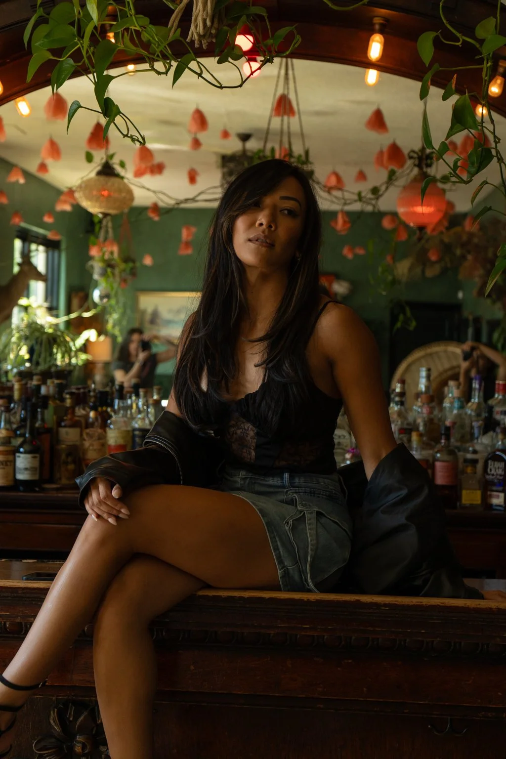 A woman with long dark hair sitting on a wooden bar counter in a dimly lit bar or restaurant, with bottles of alcohol behind her and hanging pink paper lanterns or decorations from the ceiling. Green plants and warm lighting create a cozy ambiance.