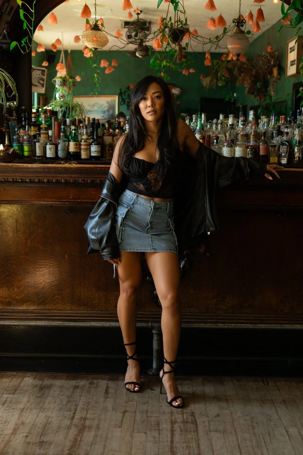 A woman standing in a bar with her arm resting on the bar counter, wearing a black top, a denim mini skirt, black high heels, and a black leather jacket draped over her shoulders. The bar workspace behind her is filled with various bottles of alcohol