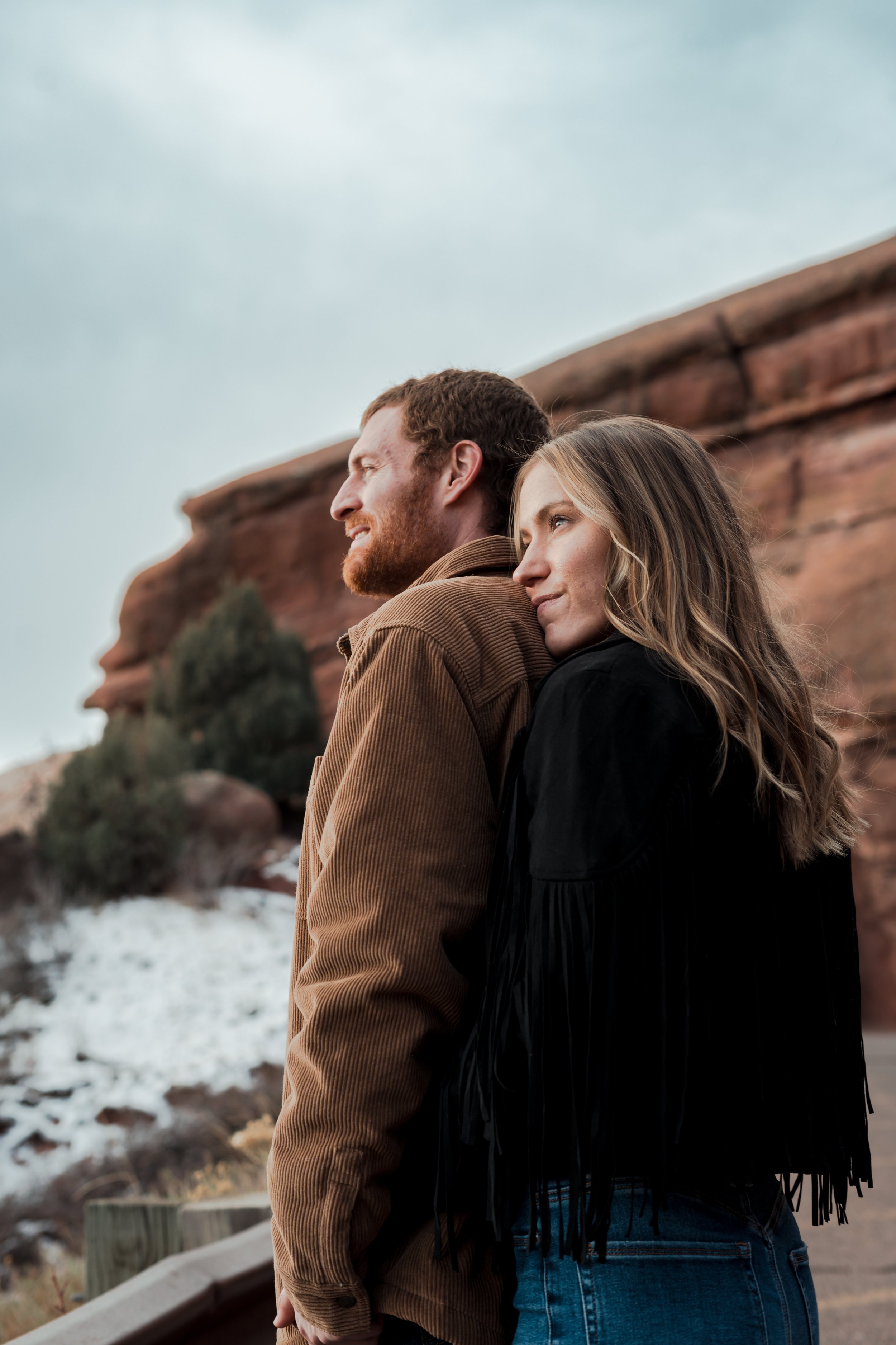 A couple stands outdoors, leaning back to each other with a rocky landscape behind them.